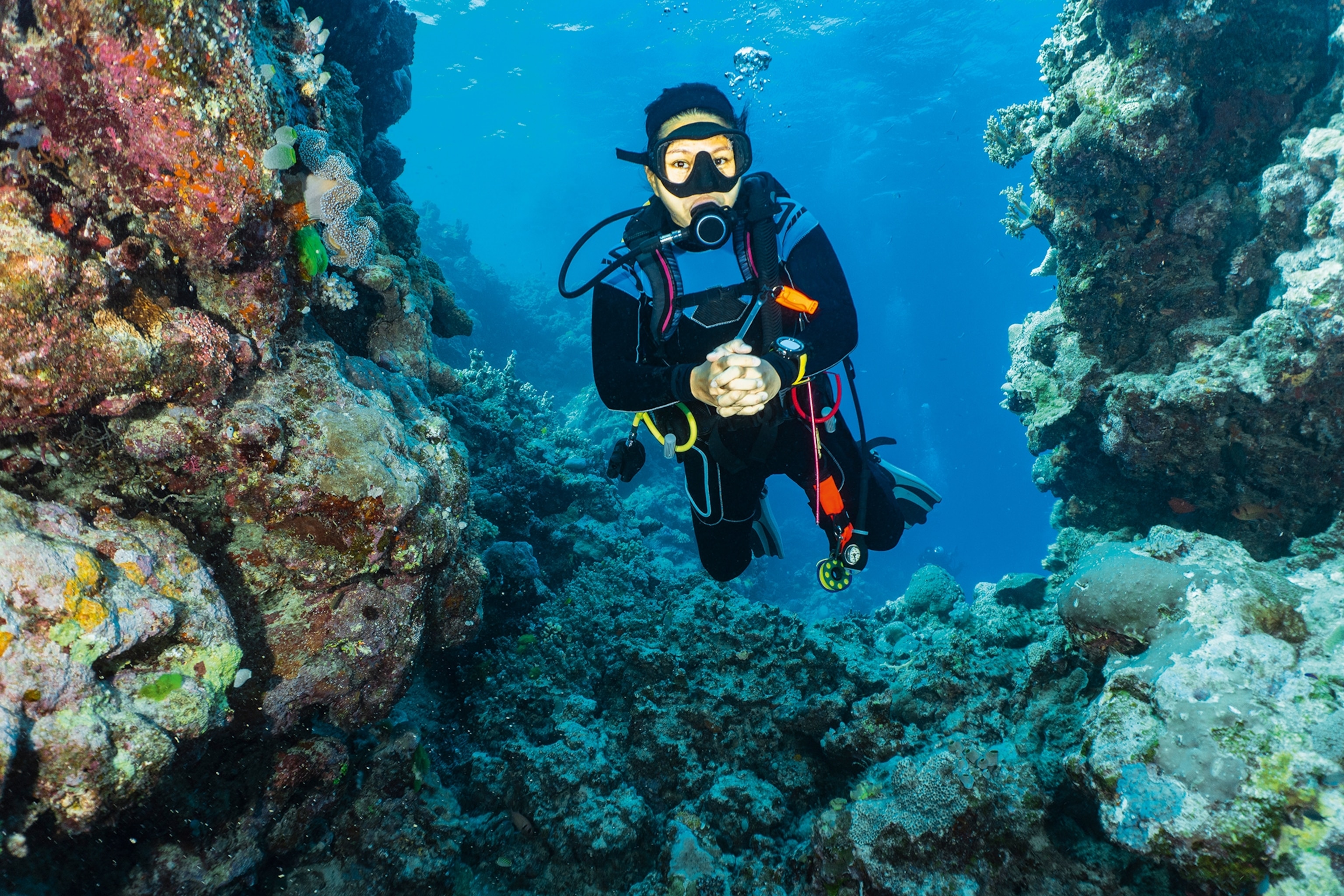 An underwater shot of a female diver showing hand signs, floating in between a cliff ridge with corals.