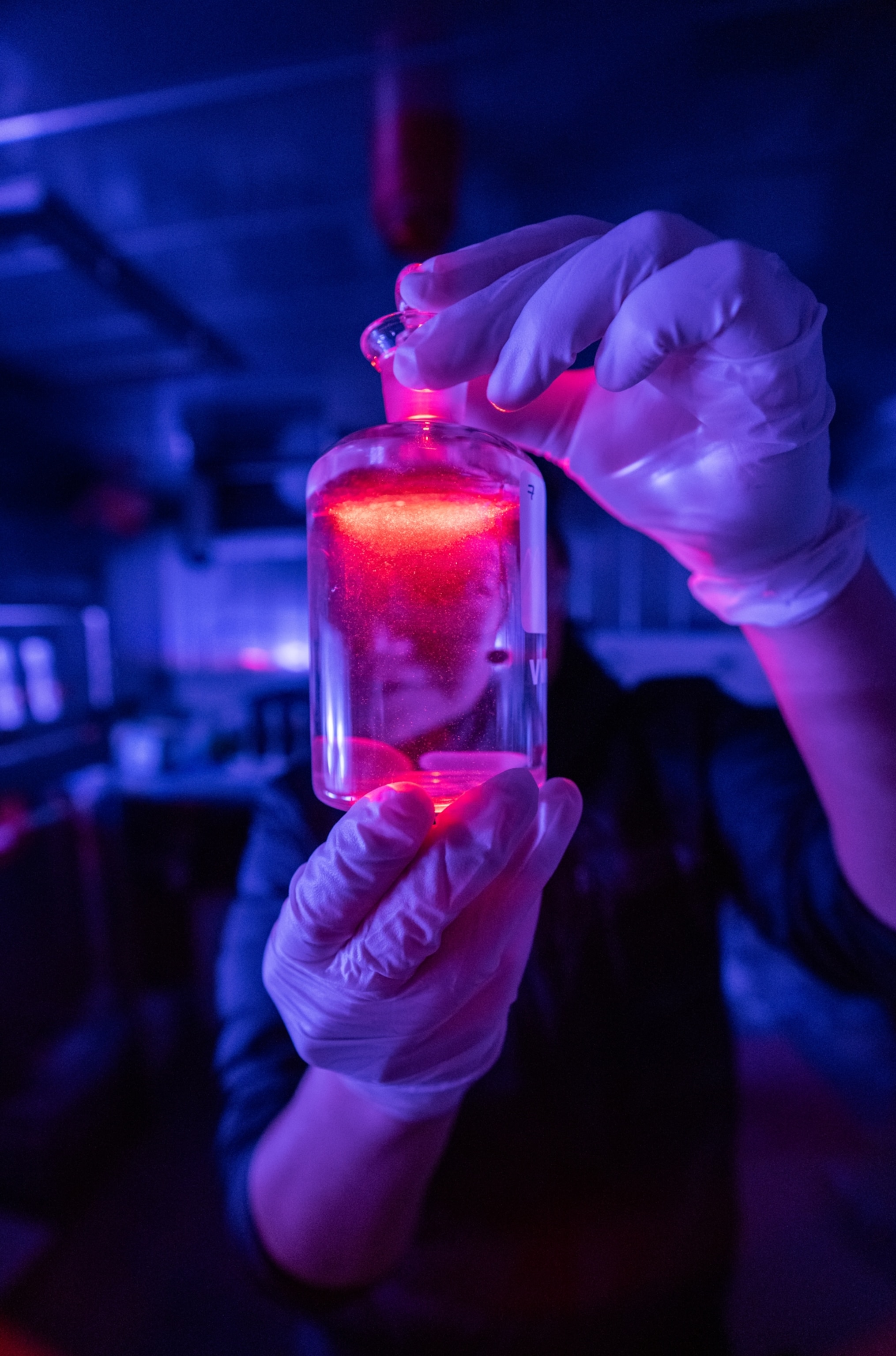 Hands in latex gloves holding glass lab bottle glowing red.