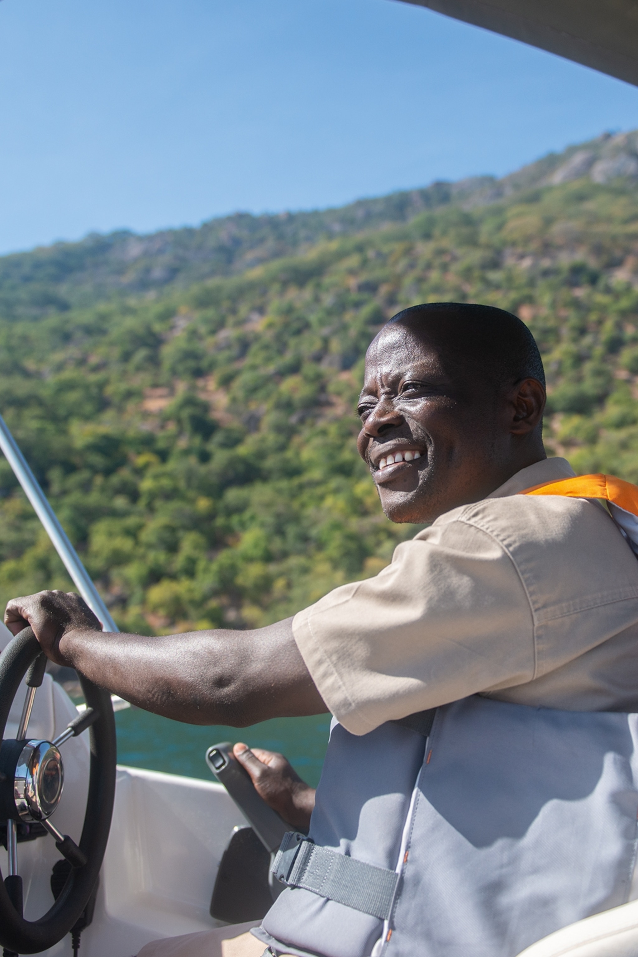 A smiling, local man steering a boat with green-carpeted hills in the background.
