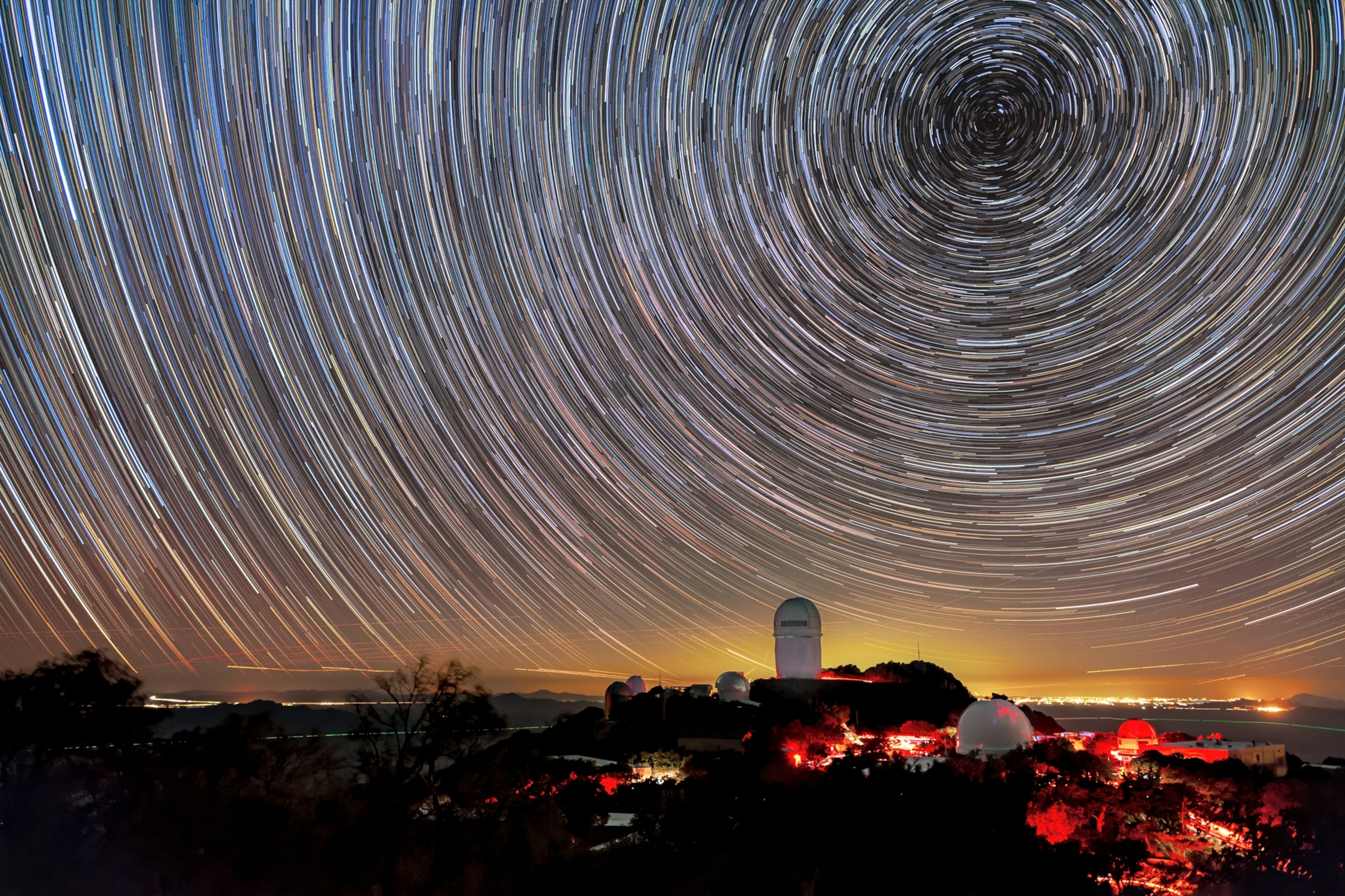 Star trails form circular patterns in the night sky over a hillside observatory with bright red lights. The horizon glows with a soft orange hue.