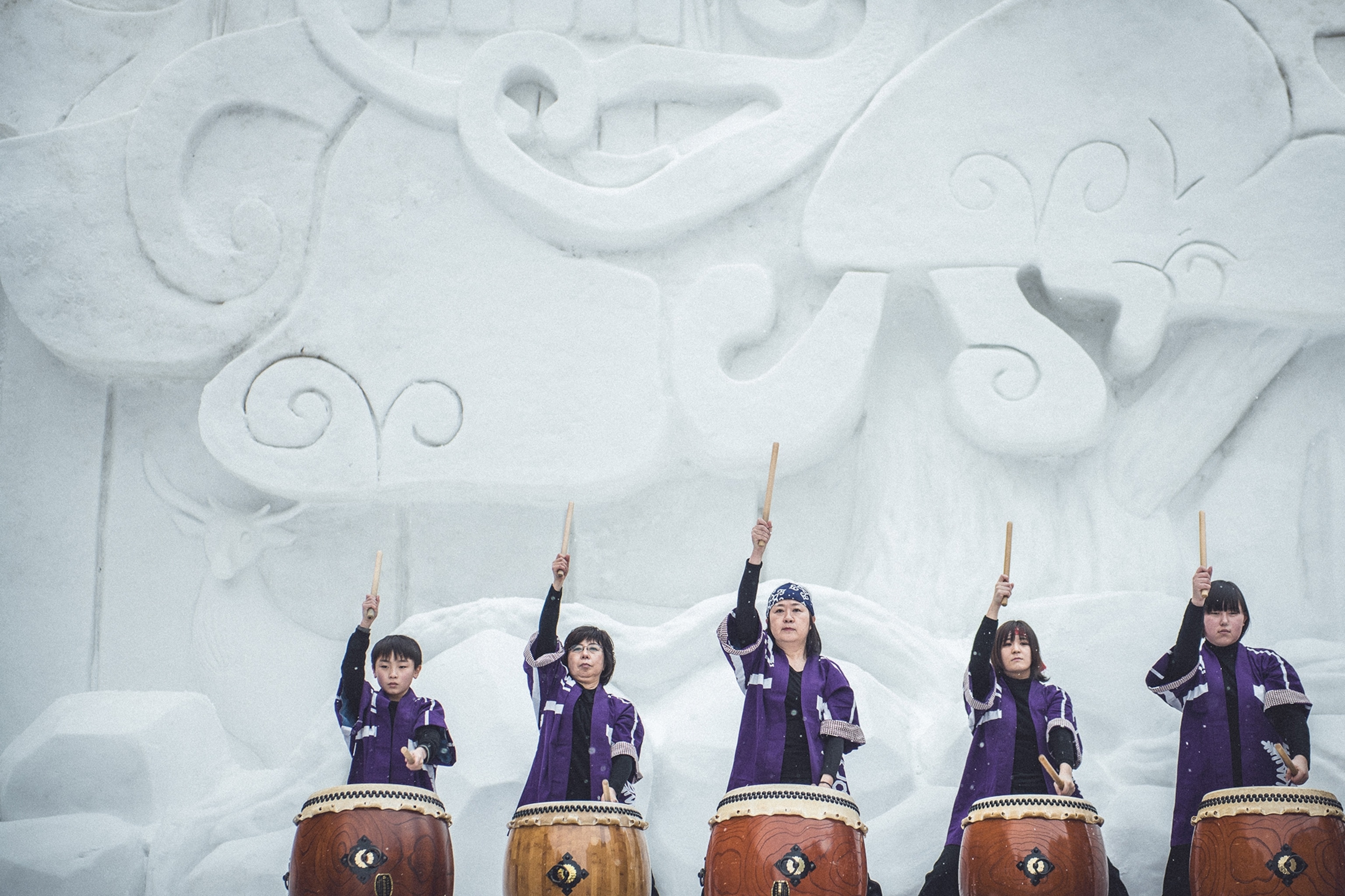 A group of indigenous, Japanese women at different ages, as well as a boy, pounding on big barrel drums in front of a sculpted ice wall.