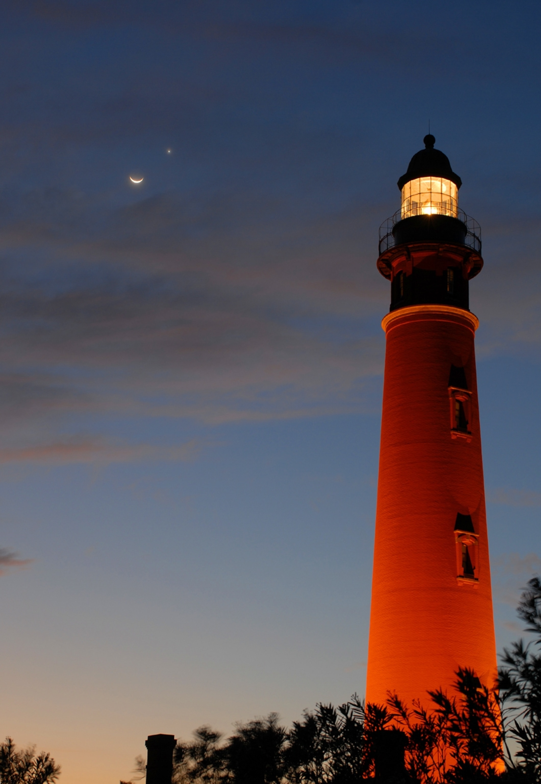 A lighthouse glows warmly at dusk, silhouetted against a deep blue sky with a crescent moon and Venus visible close together