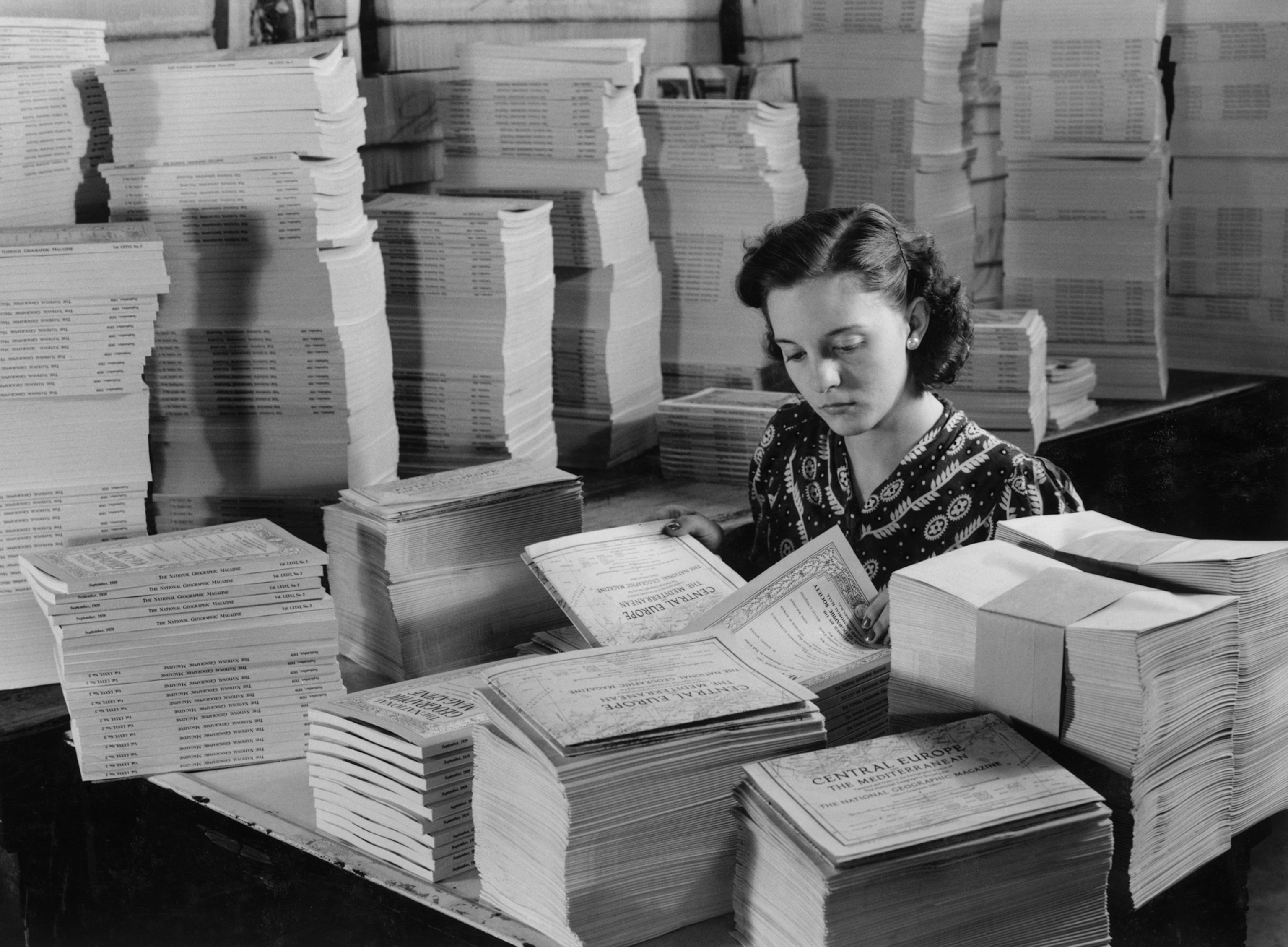 business A young woman is surrounded by stacks of magazines and is inserting papers into them