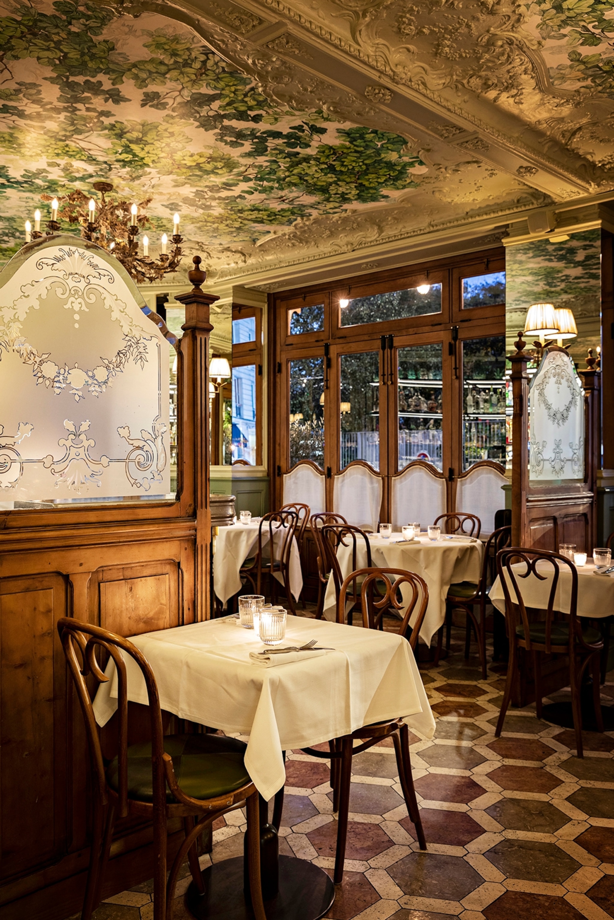 The interior of a Belle Epoque French cafe, with wooden bistro chairs, a tile floor and a fresco ceiling.