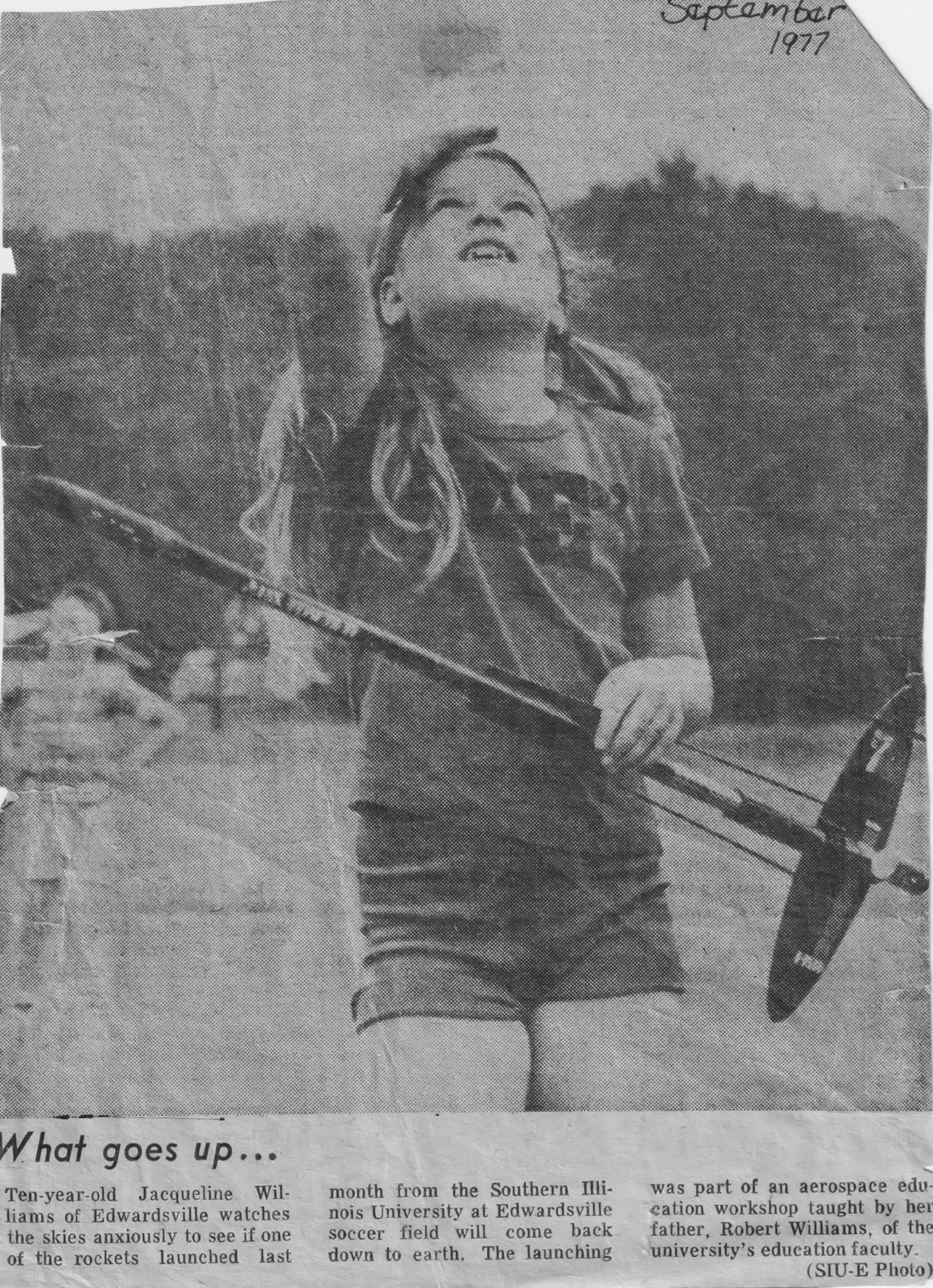 A young girl with a ponytail eagerly looks up at the sky, holding a small model rocket. It's a black-and-white newspaper image from September 1977, conveying curiosity.