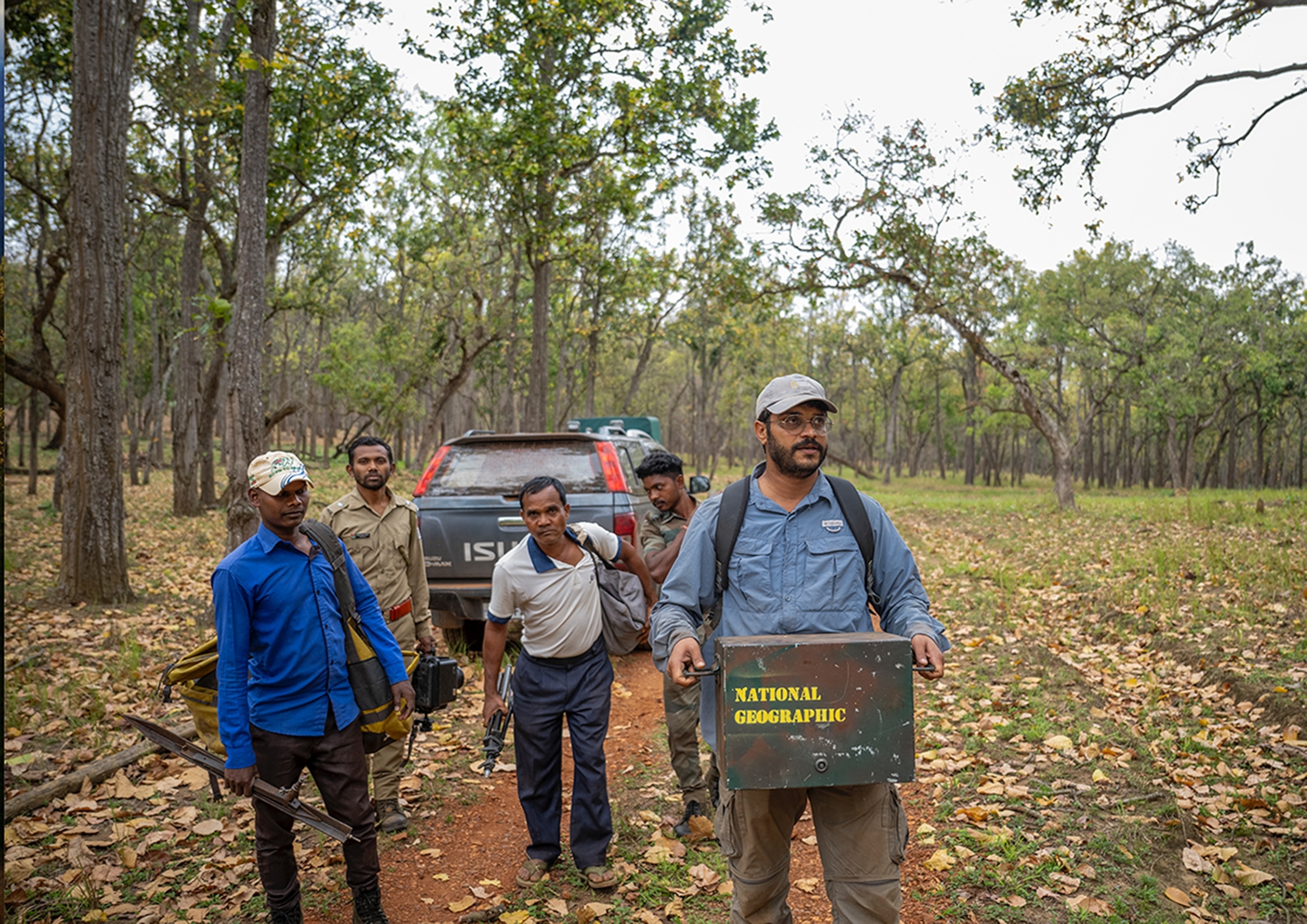 A group of people walk in the woods. One of them is holding a wooden box that reads 'National Geographic'