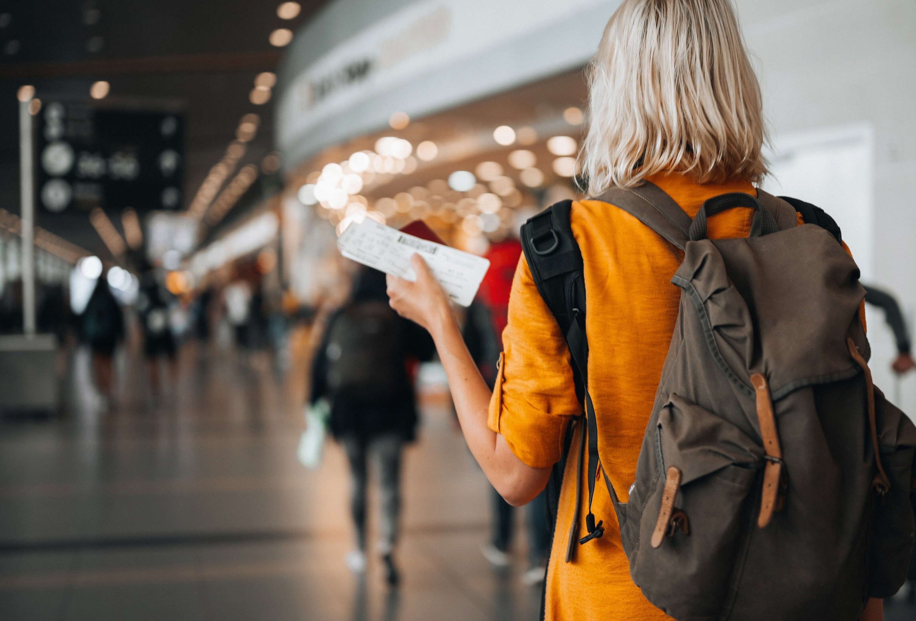 A woman wearing an orange top holds a boarding pass in her left hand as she walks through an airport.