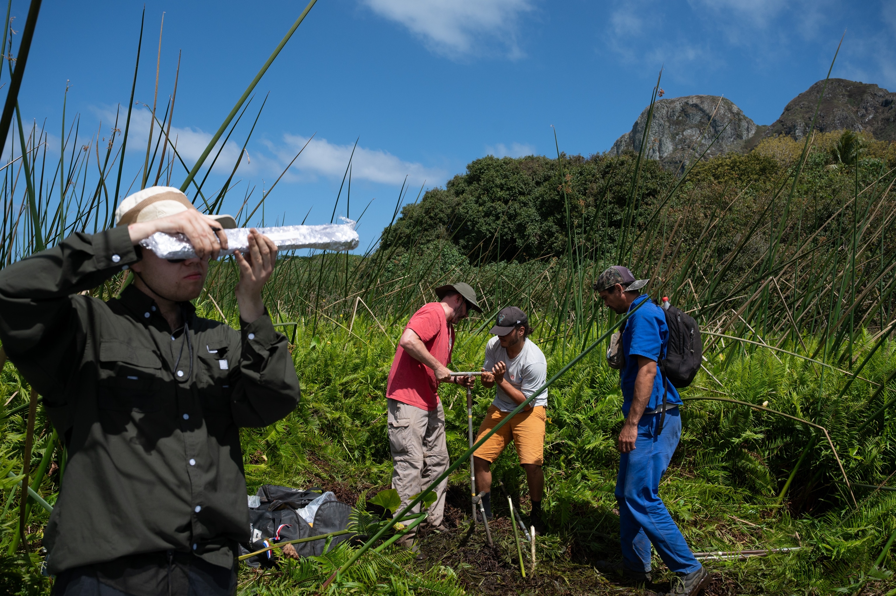 A person inspects a wrapped up core sample in the foreground as two other people in the background pull another core sample as another person watches.