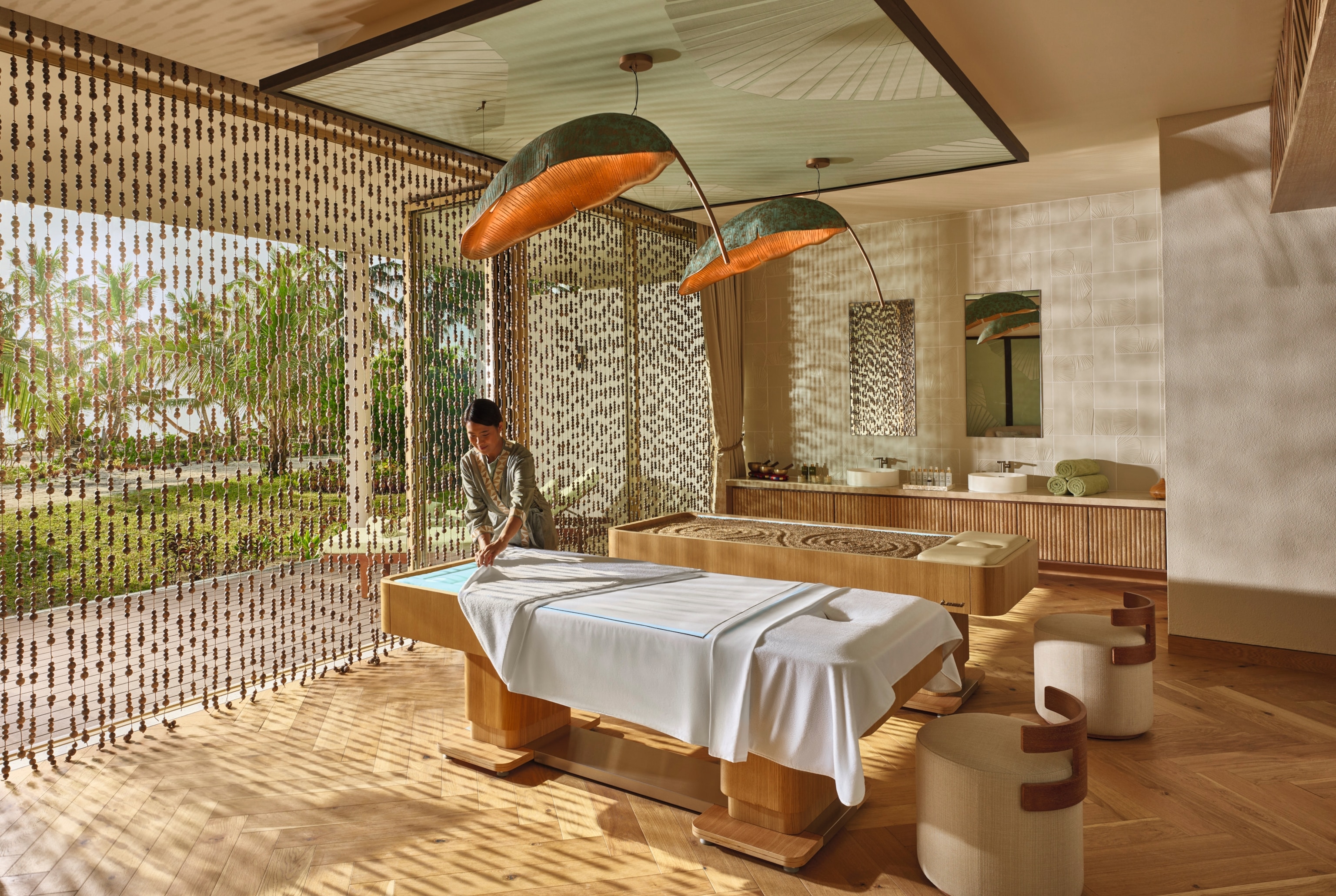 Sunlit luxury spa treatment room with a massage table, wooden interiors, and beaded curtains overlooking a garden full of palm trees. 



