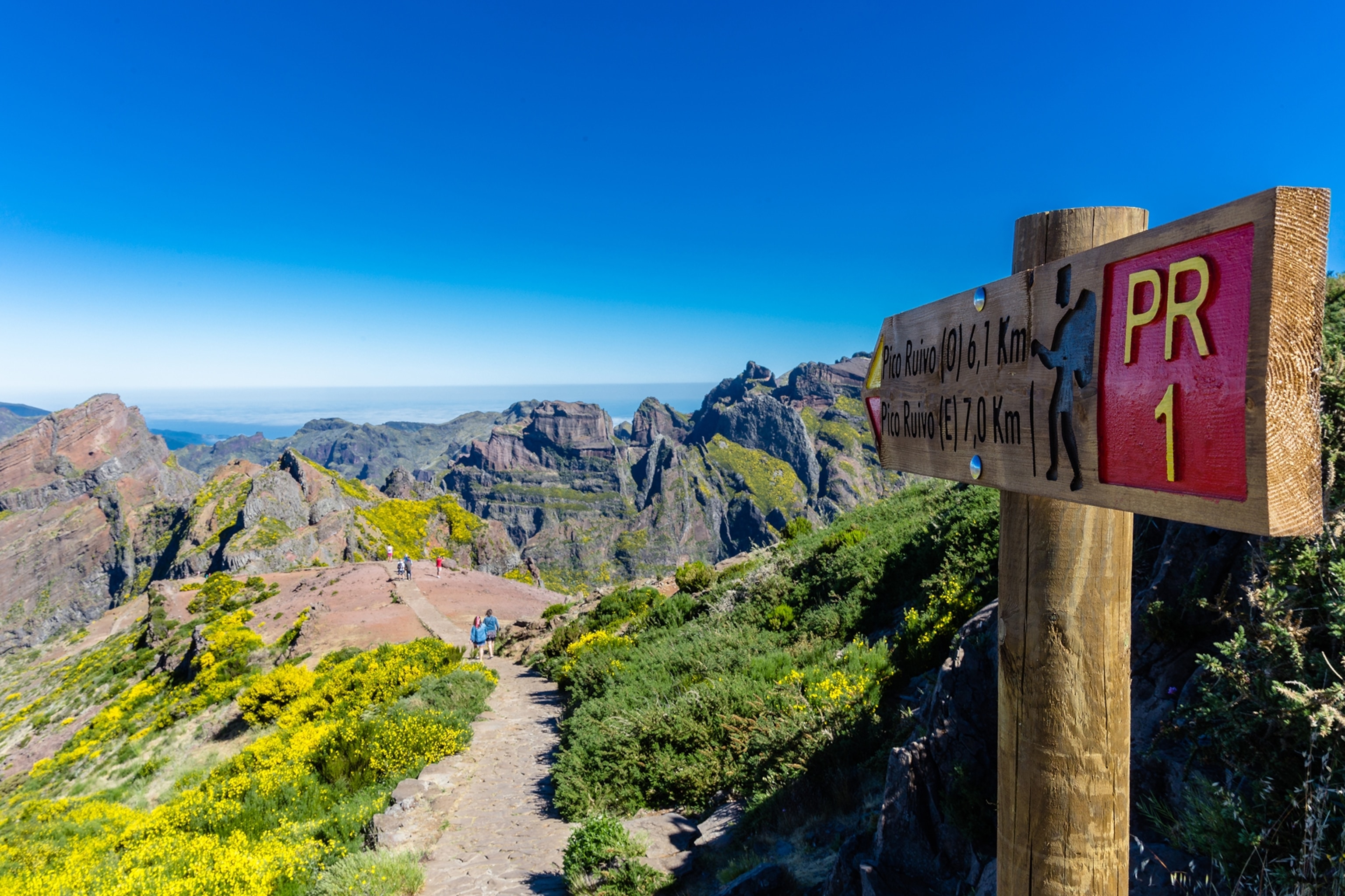 A trail leading along the ridge of a mountain with a sign post pointing in its direction, mountains are rising in the distance.