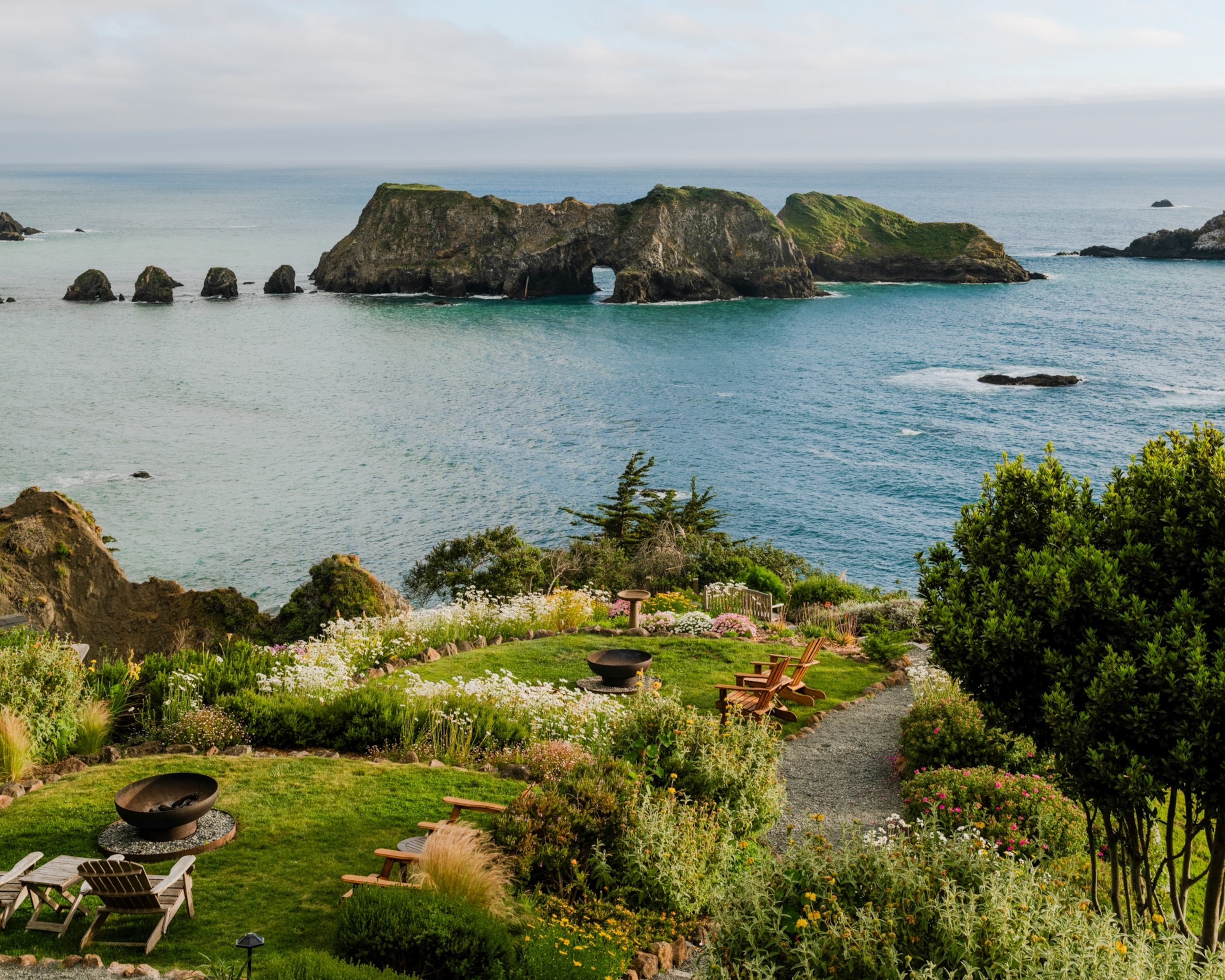 A landscaped garden with seating areas overlooks the ocean, with a rocky island featuring a natural arch rising from the water offshore.
