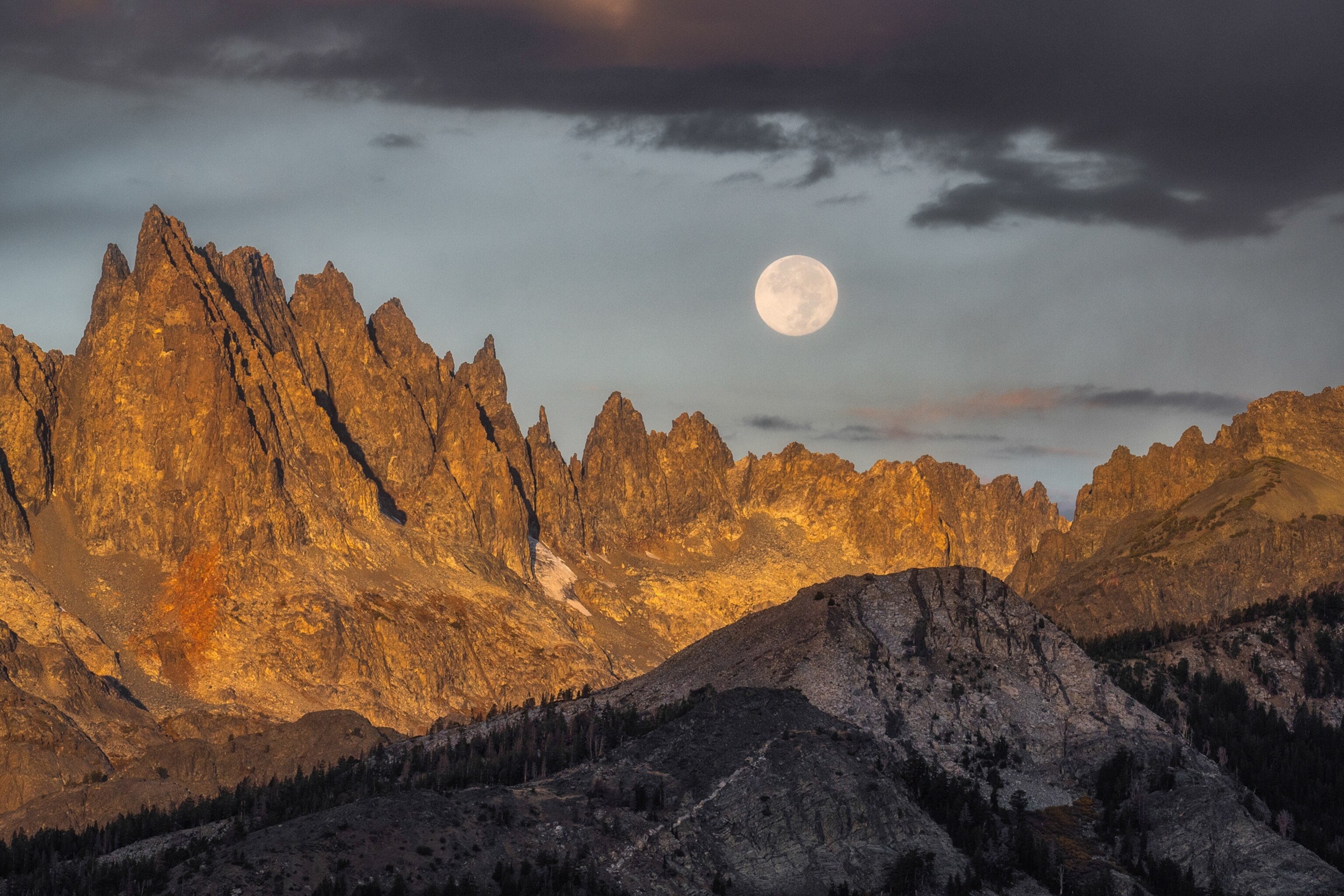 A full moon rises above a rugged mountain range, illuminating sharp rocky peaks beneath dark clouds in the evening sky.