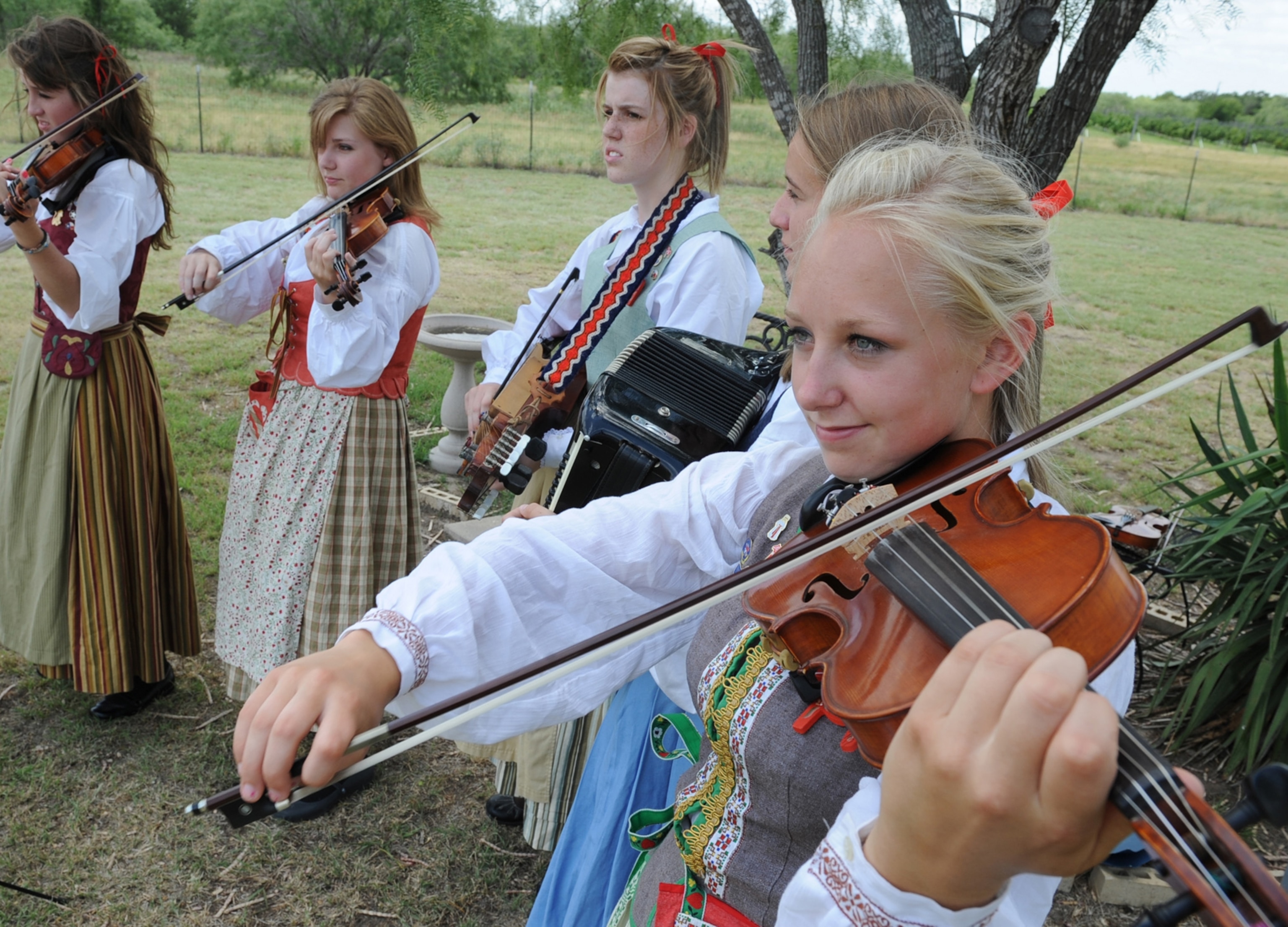 Five young women in traditional folk costumes play violins and an accordion outdoors