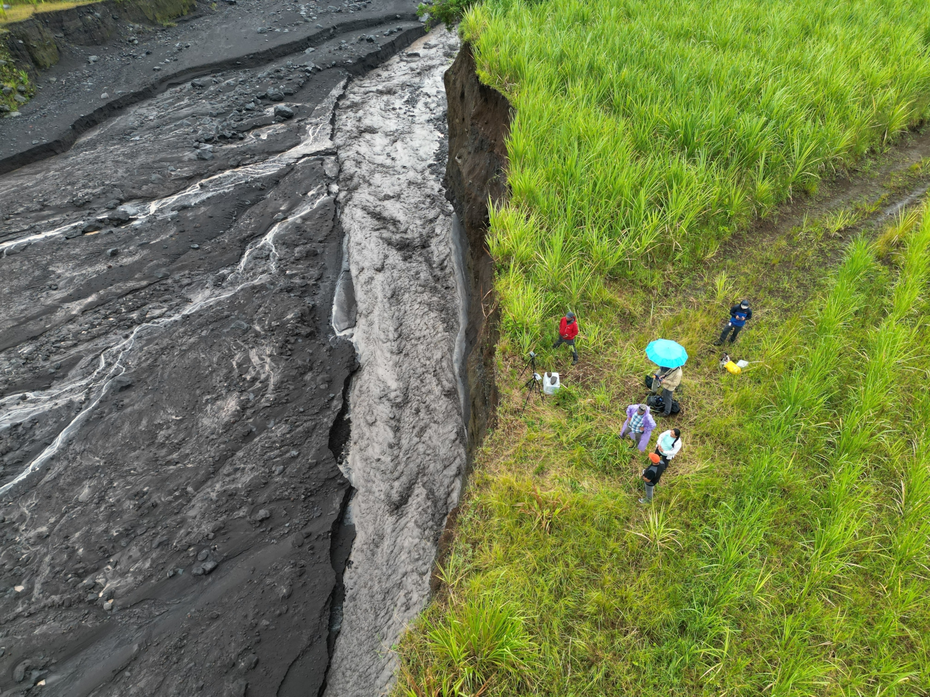 Aerial view of people standing on vibrant green grass near a sharp cliff edge overlooking a dry lava flow.
