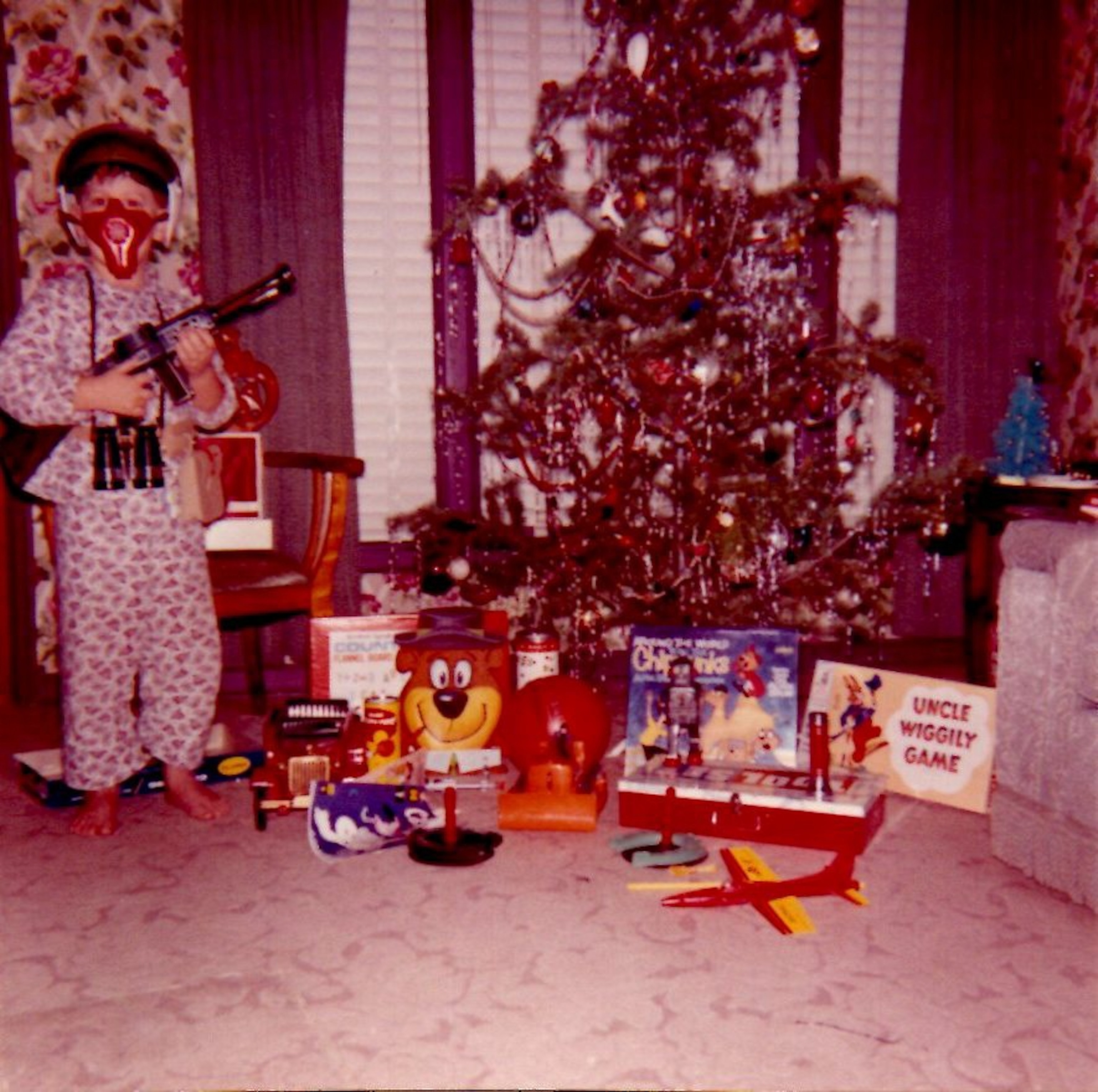 A child in pajamas, wearing a rocket helmet, holds a toy gun by a decorated Christmas tree.