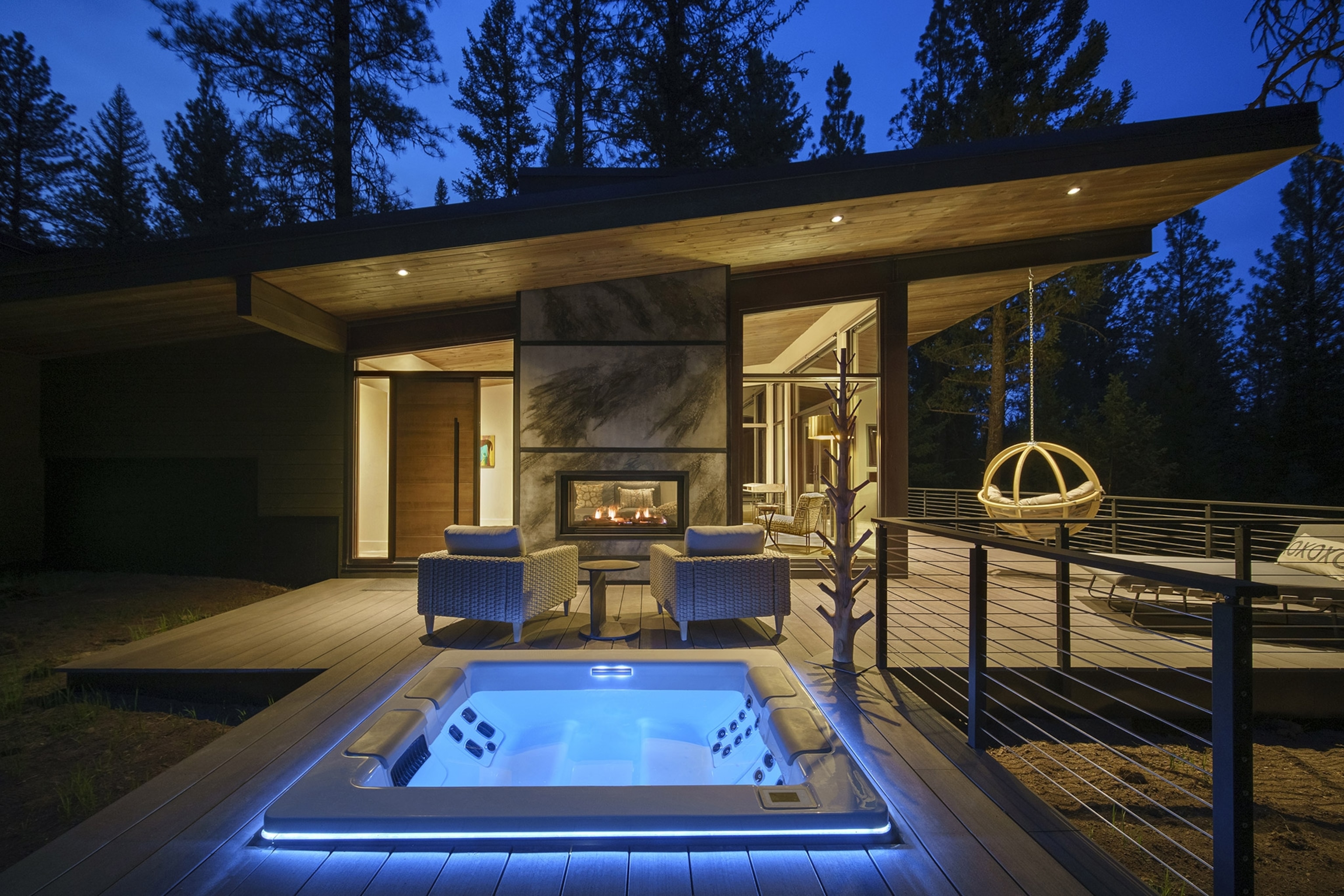 A square hot tub with blue lights in the foreground, set into a deck extending out from a small warmly lit cabin. A darkening sky above and silhouettes of trees behind.