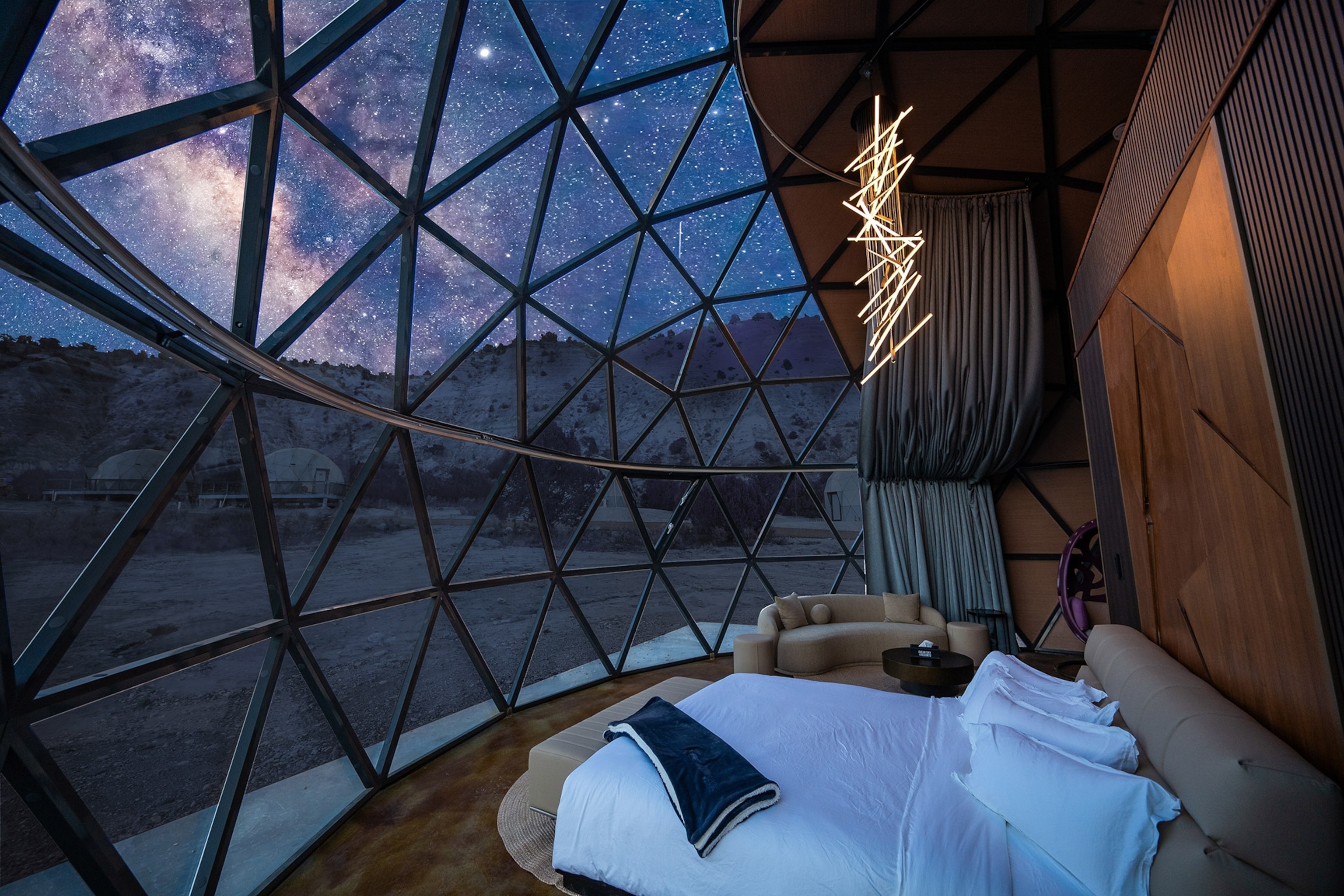 Interior of a glass dome with geometric pattern on the glass. A bed with white bedding facing outward, the sky showing brilliant stars