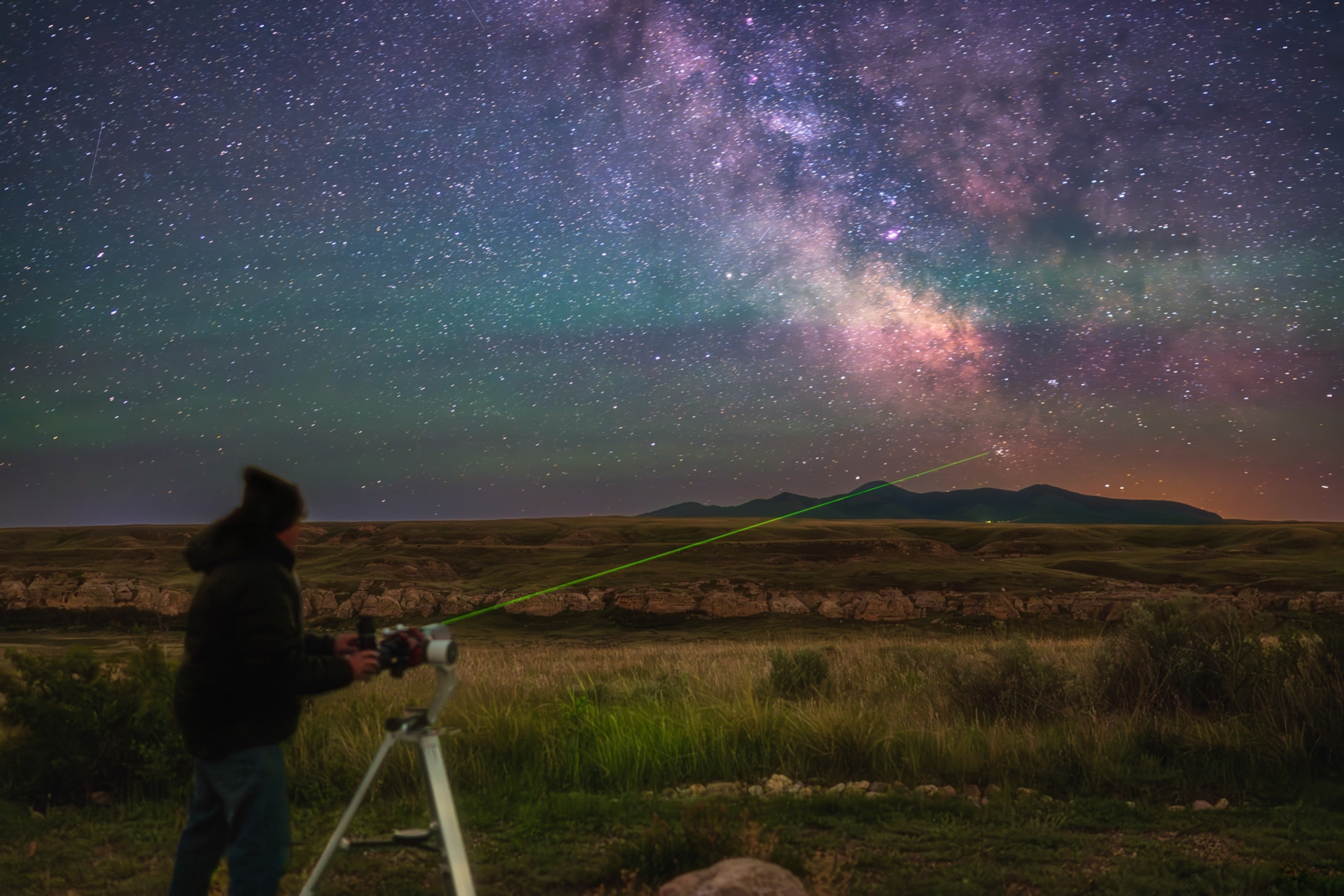 An astronomer standing in the foreground using a telescope under a vibrant night sky, the Milky Way prominently visible. The astronomer is pointing a green laser points towards the Milky Way.