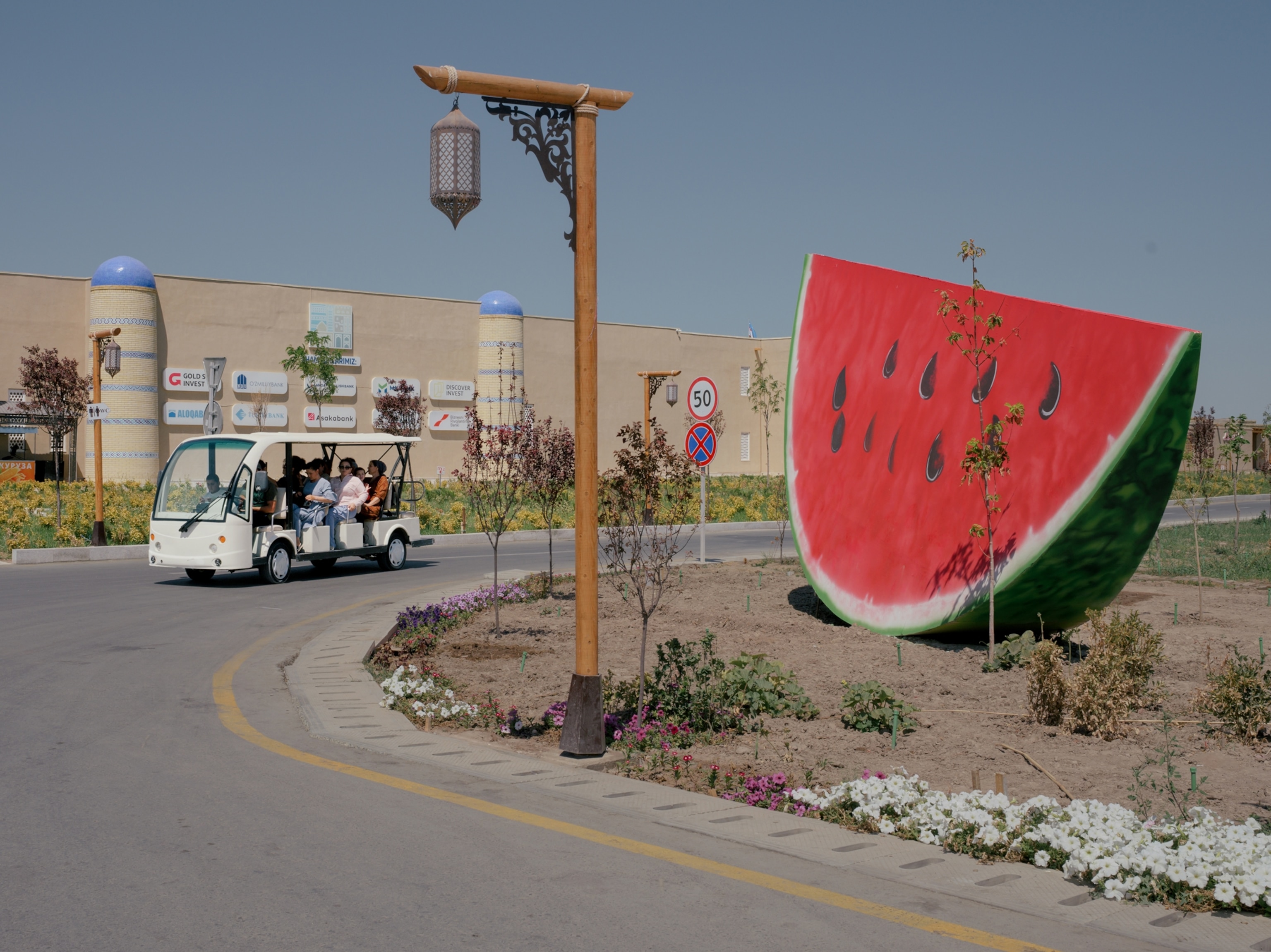 A group rides in a cart down a paved road. On the road, closer to the camera and in the direction the cart is headed, is a large watermelon sculpture painted with the red body and green rind.
