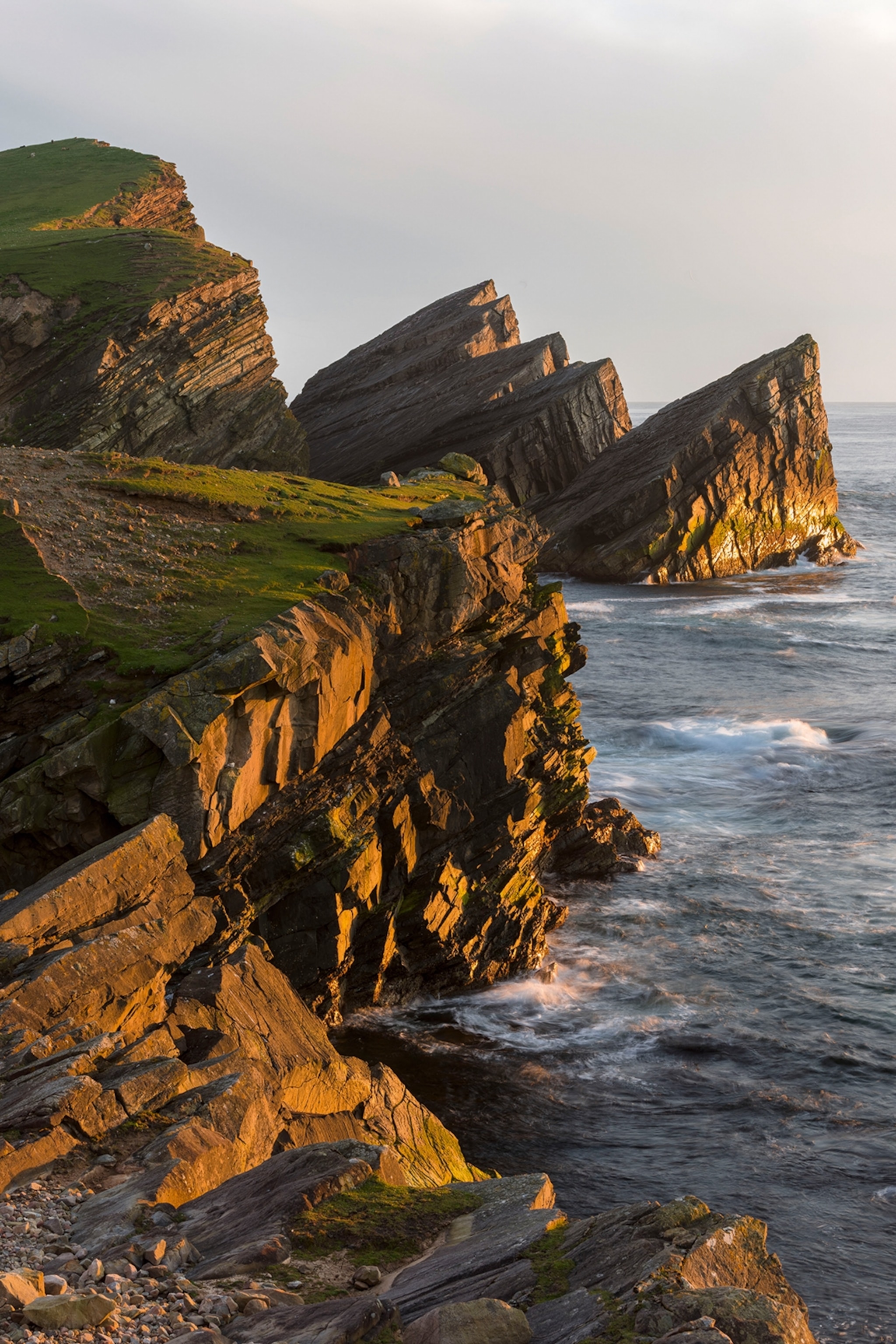A stretch of jagged sea cliffs washed in dusk sunlight as water breaks below.