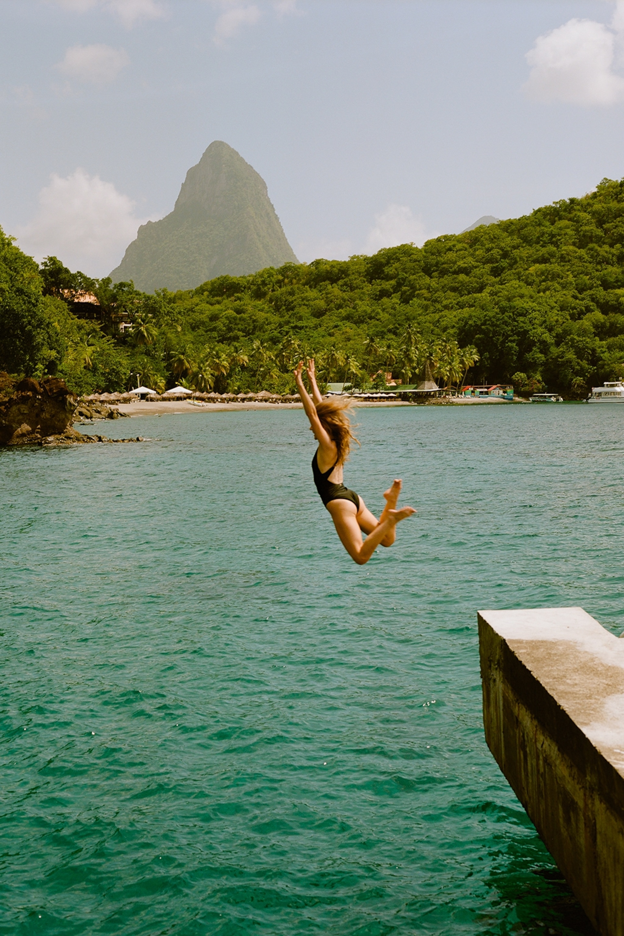 A teenage girl jumping into the ocean from a pier overlooking the shore of a Caribbean island.