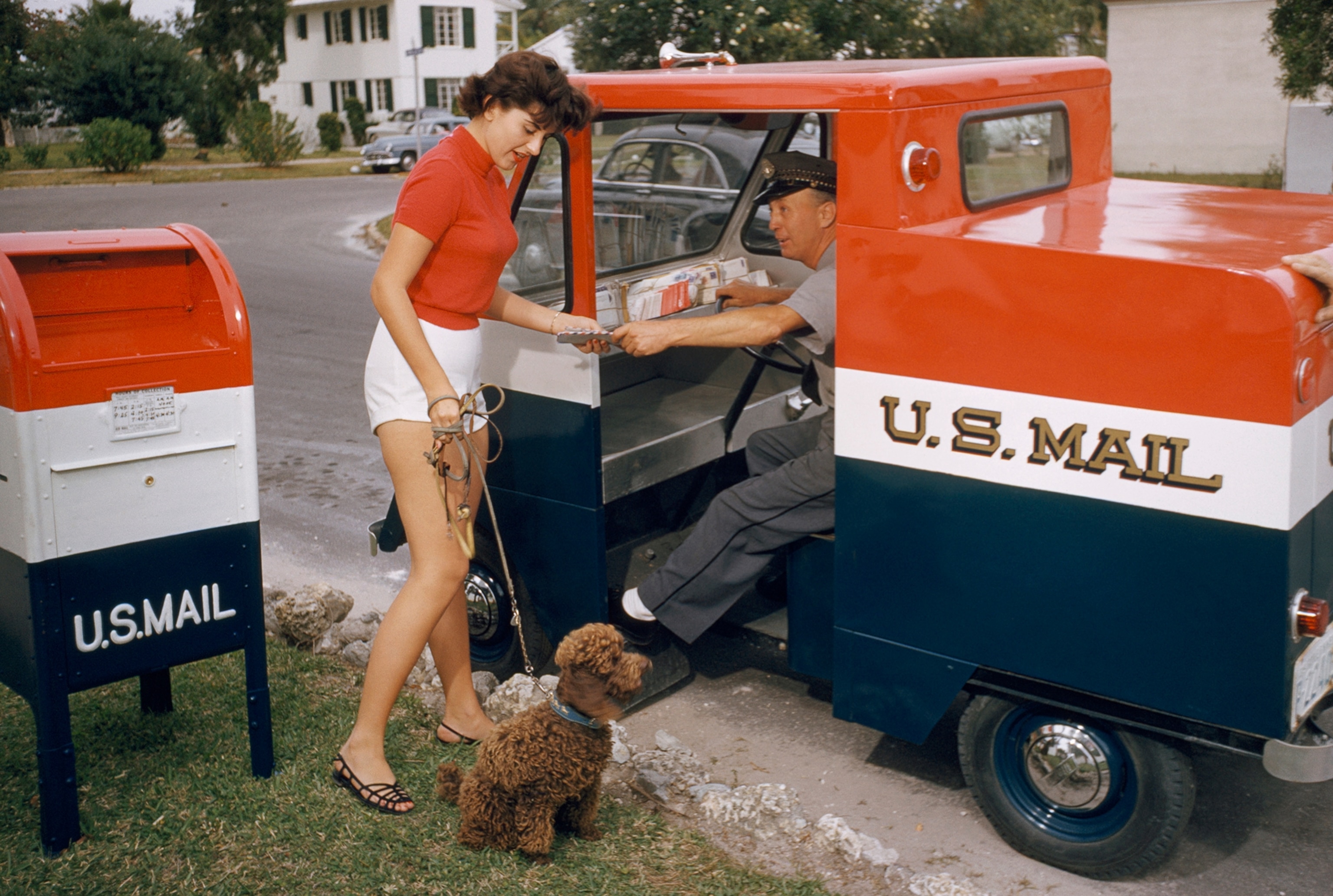 A mailman in a truck hands a letter to a girl with a dog by a mailbox.