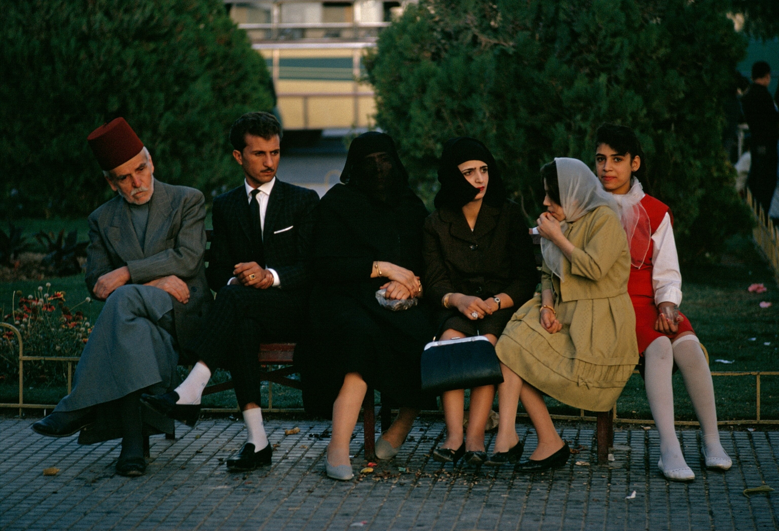 Men and women, one in a fez, sit on a park bench, 1965