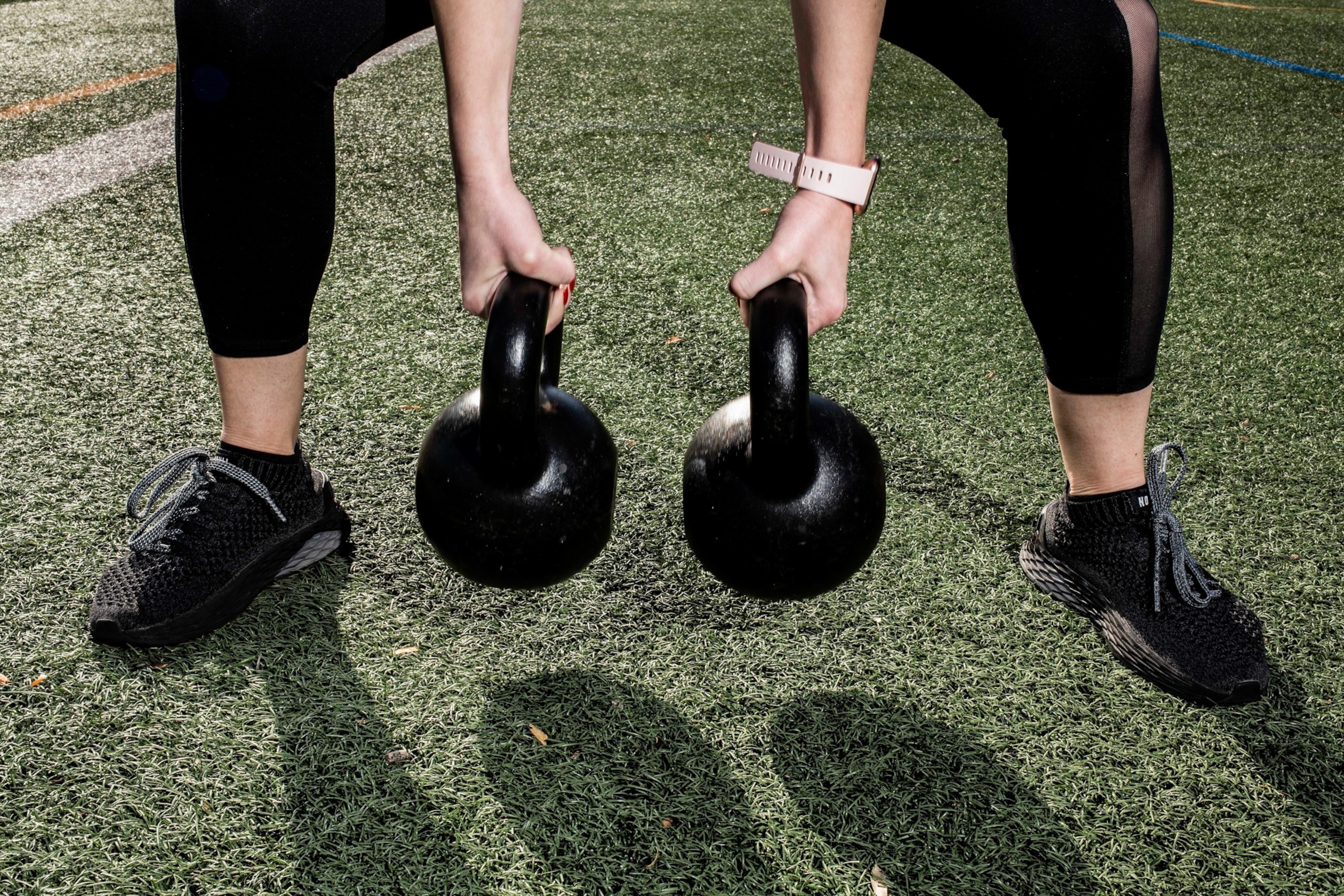 A person wearing black athletic shoes and leggings holds two kettlebells over a grassy field.