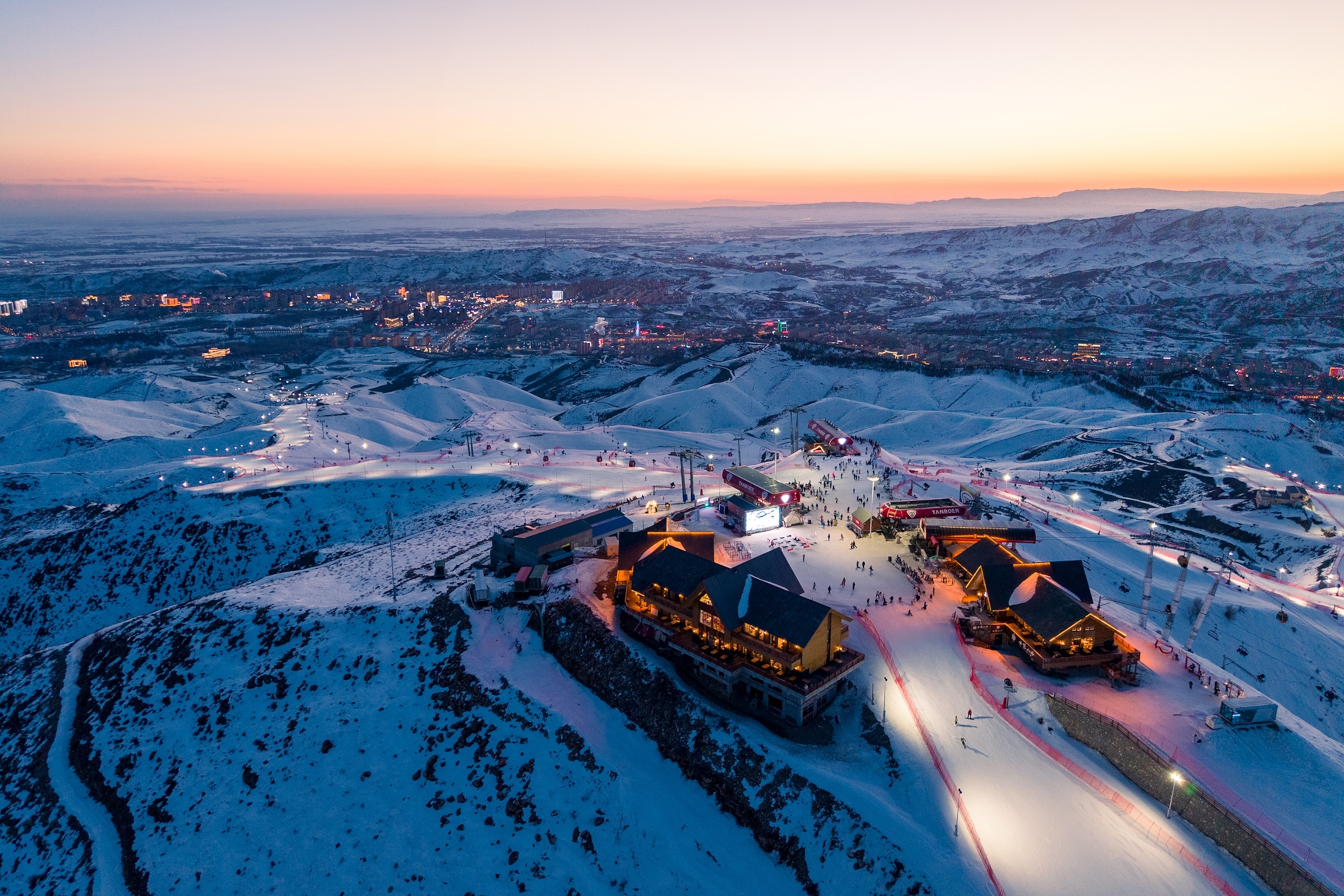 An areal view over a snowy ski resort at night with houses illuminating late dusk.