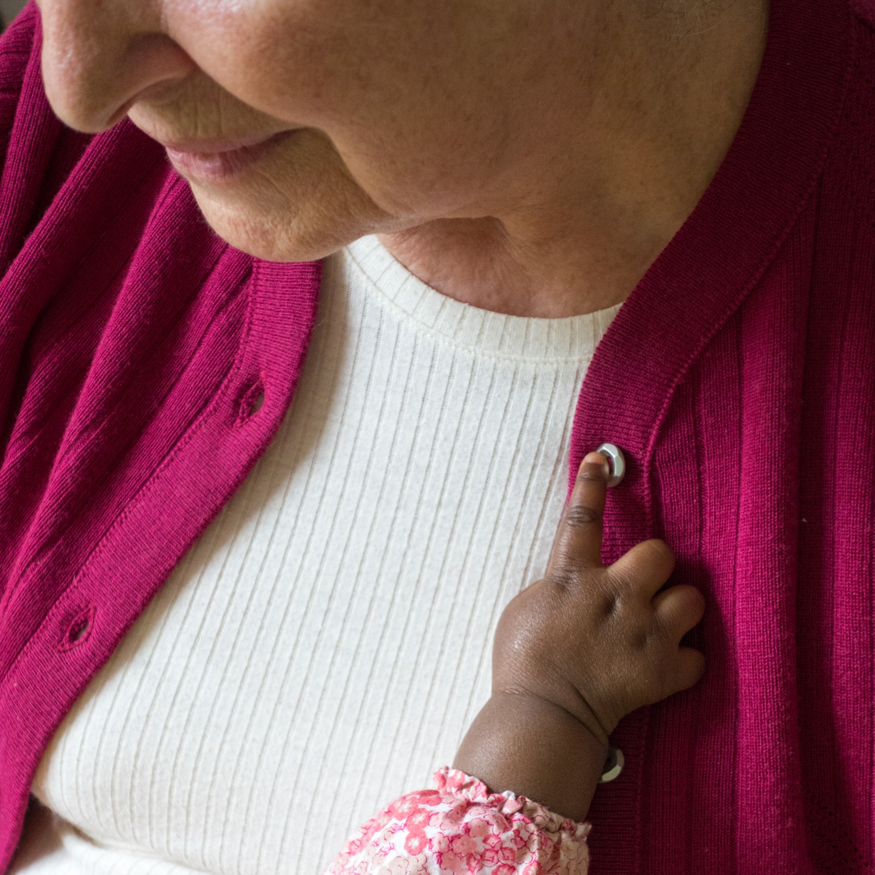 Close-up of an elderly woman in a pink cardigan, her face partially visible. A small child’s hand in a floral sleeve touches her button