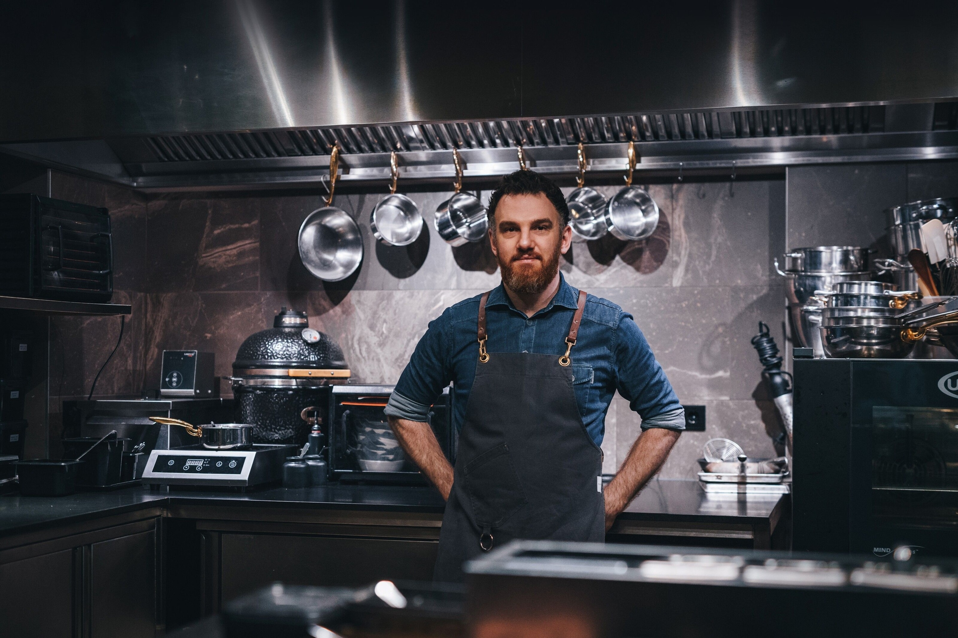 René Frank stood in the CODA kitchen. Above his blue denim shirt, he wears a plain black apron with brown leather straps.