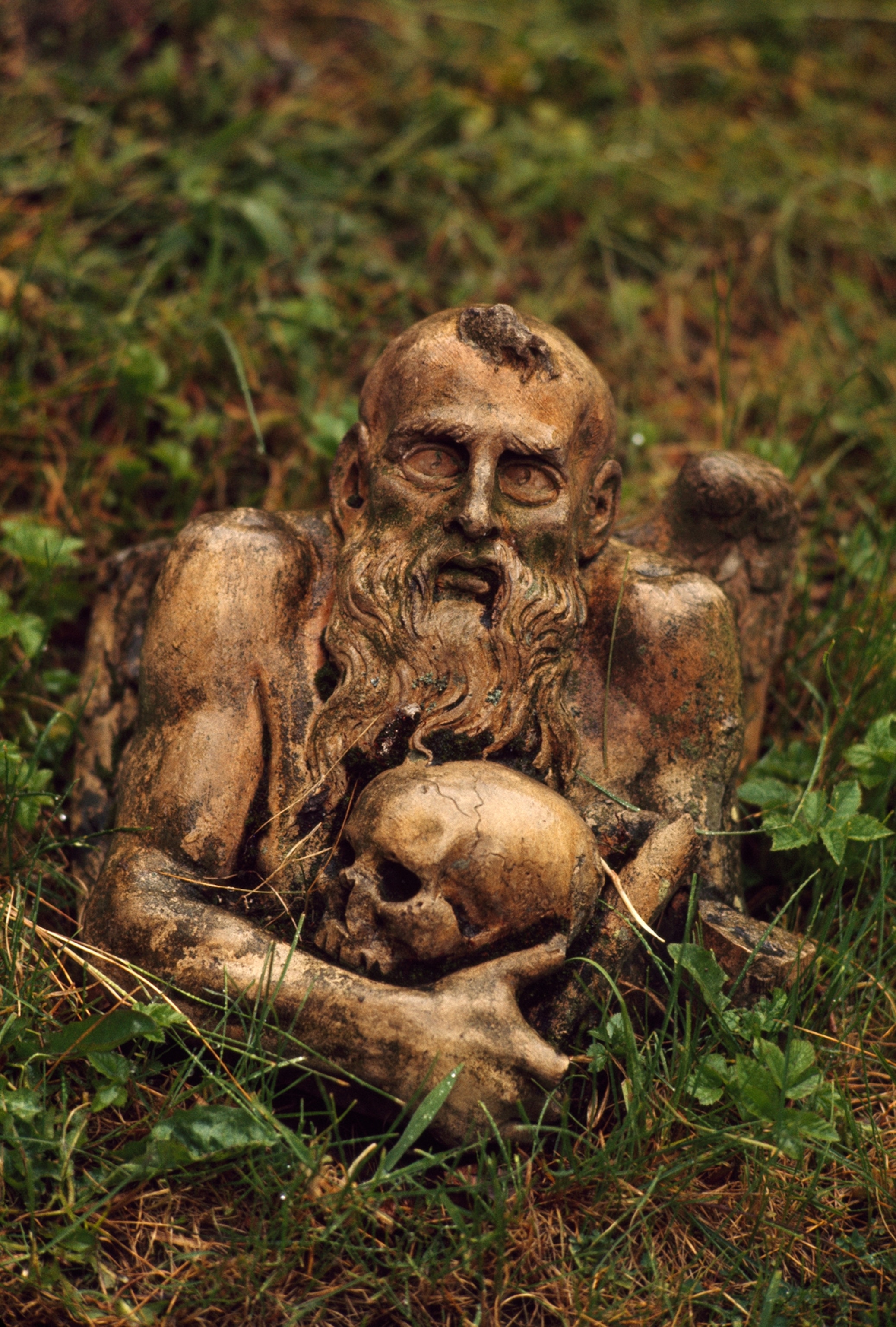 Stone carving of a bearded man cradling a skull lies in a field.