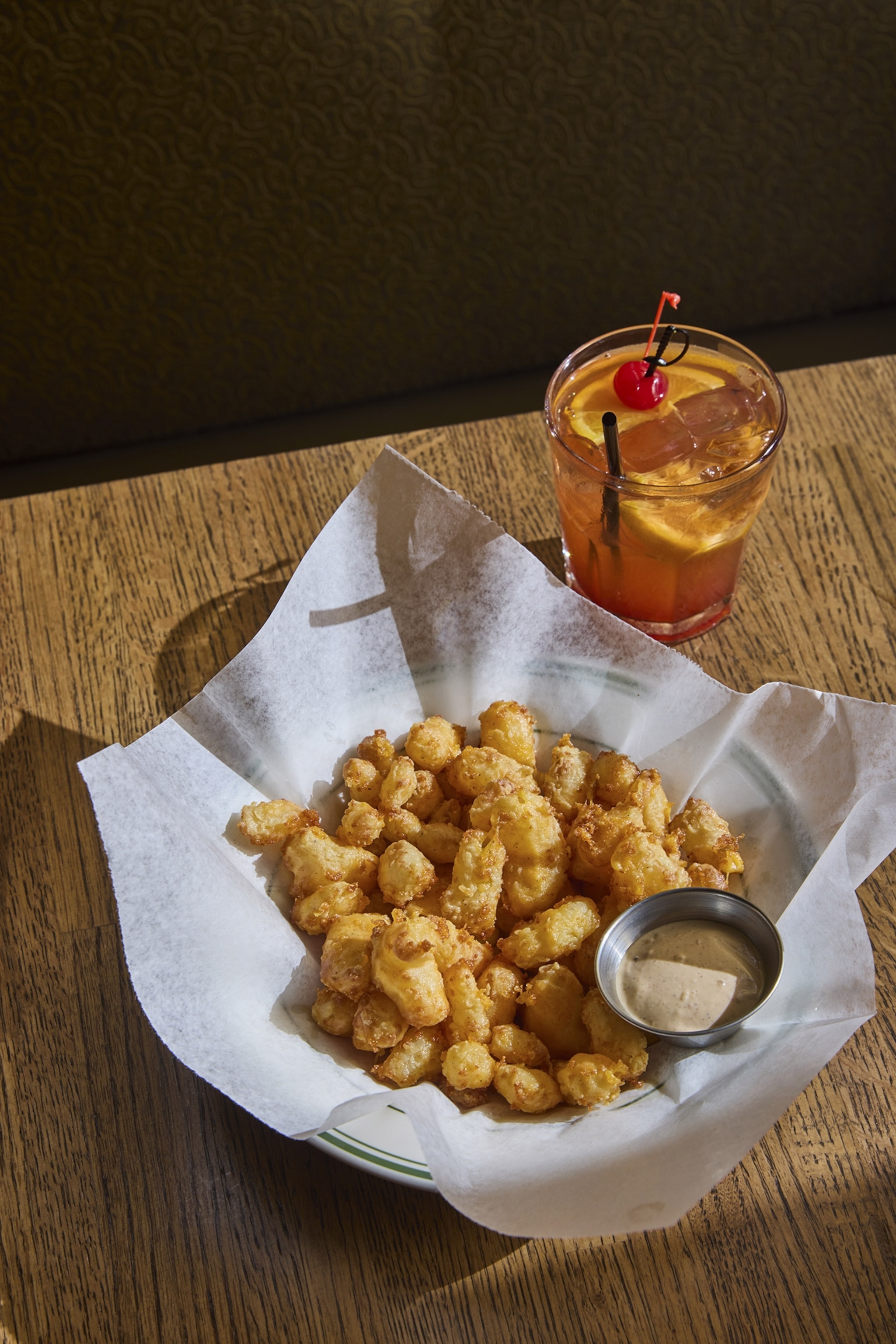 A paper-lined basket contains fried cheese curds and dipping sauce, accompanied by a cocktail in a short glass with a cherry and orange garnish