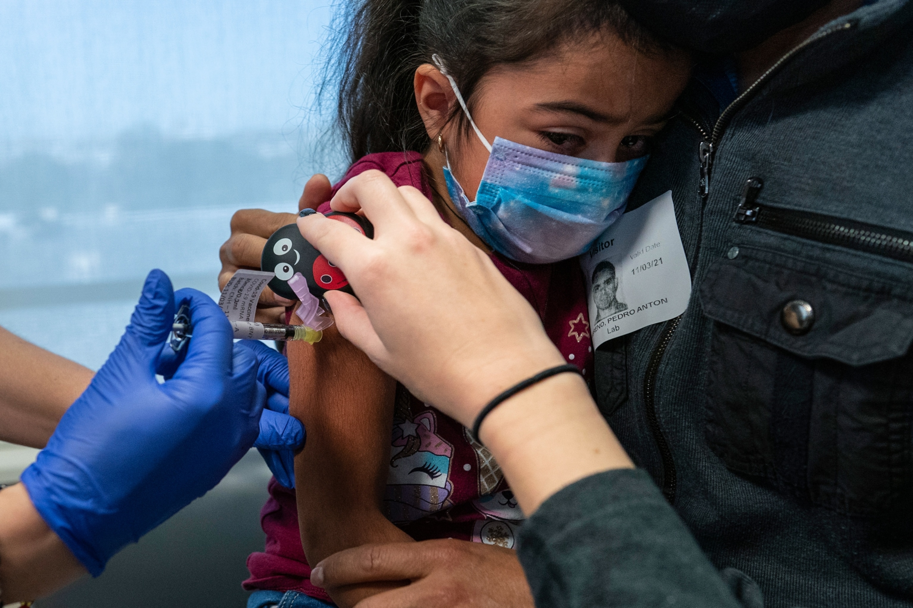 A young girl wearing a mask held in her father's arms as a nurse holds a device on her arm while administering a vaccine injection below it.
