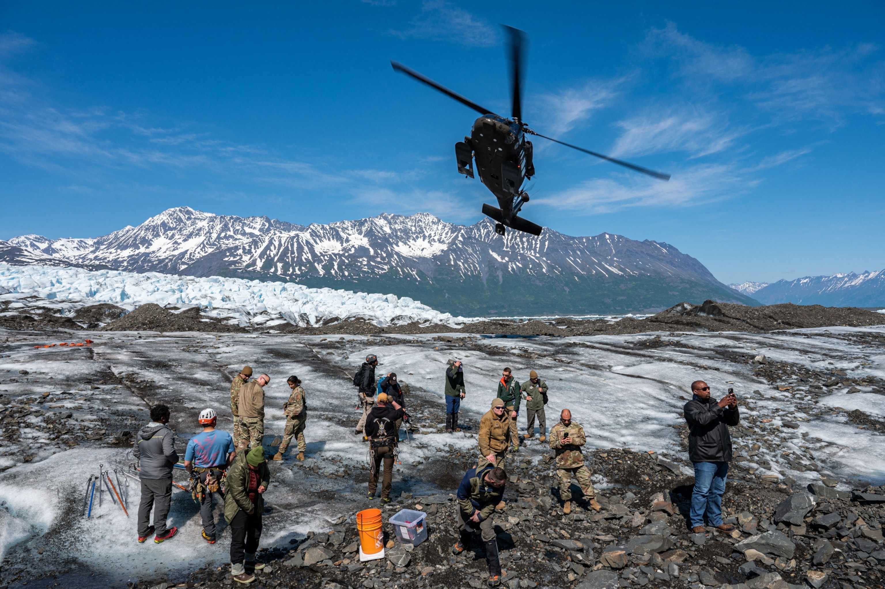 Helicopter hovers over a glacier with snow-capped mountains in the background. A group of people in outdoor gear stand on the ice