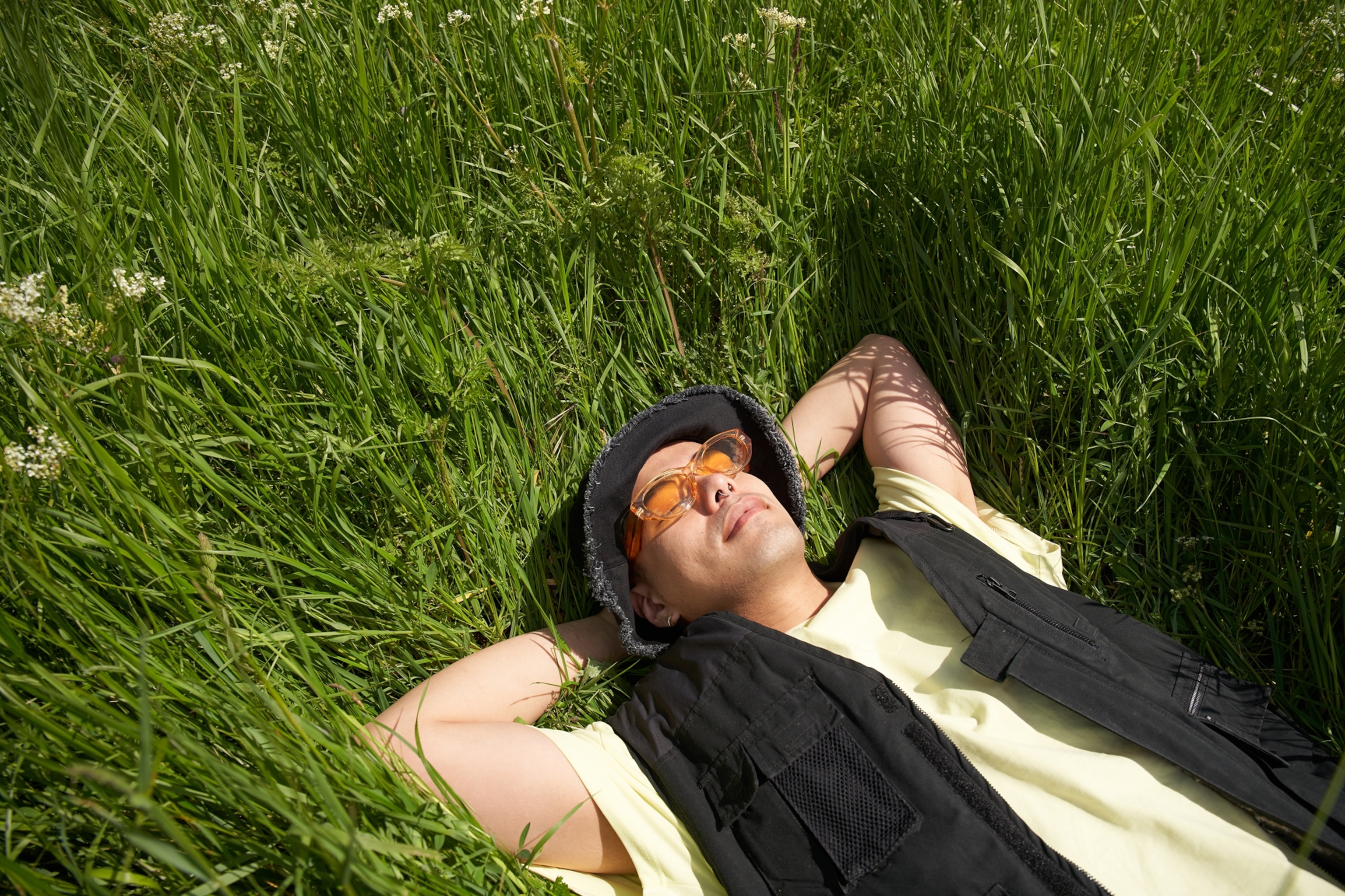 A man wearing a black bucket hat, orange sunglasses, and a yellow shirt with a black vest lies on his back in a field of grass.
