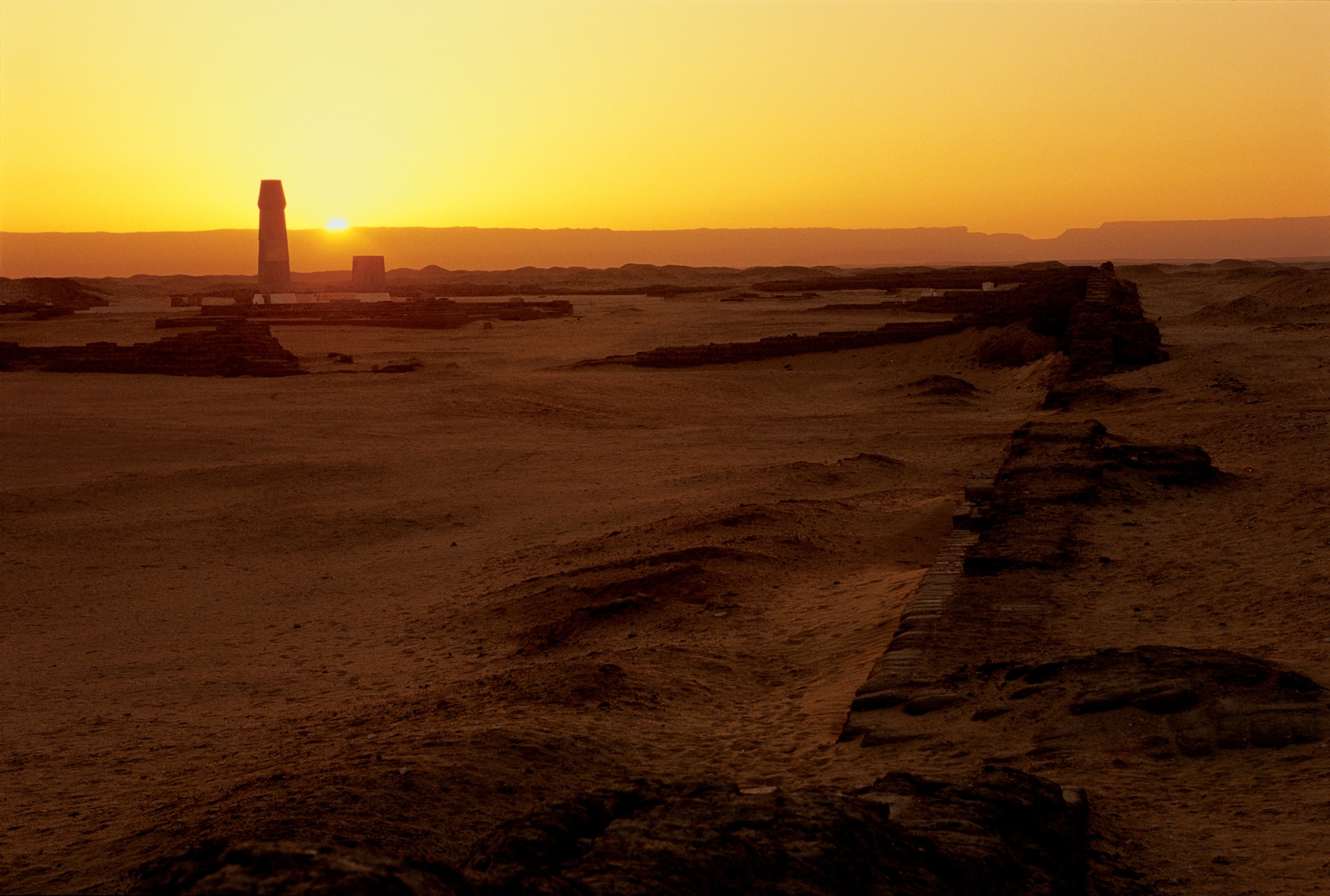 A vast desert landscape at sunset, featuring ancient ruins and a tall, narrow monument silhouetted against an orange sky