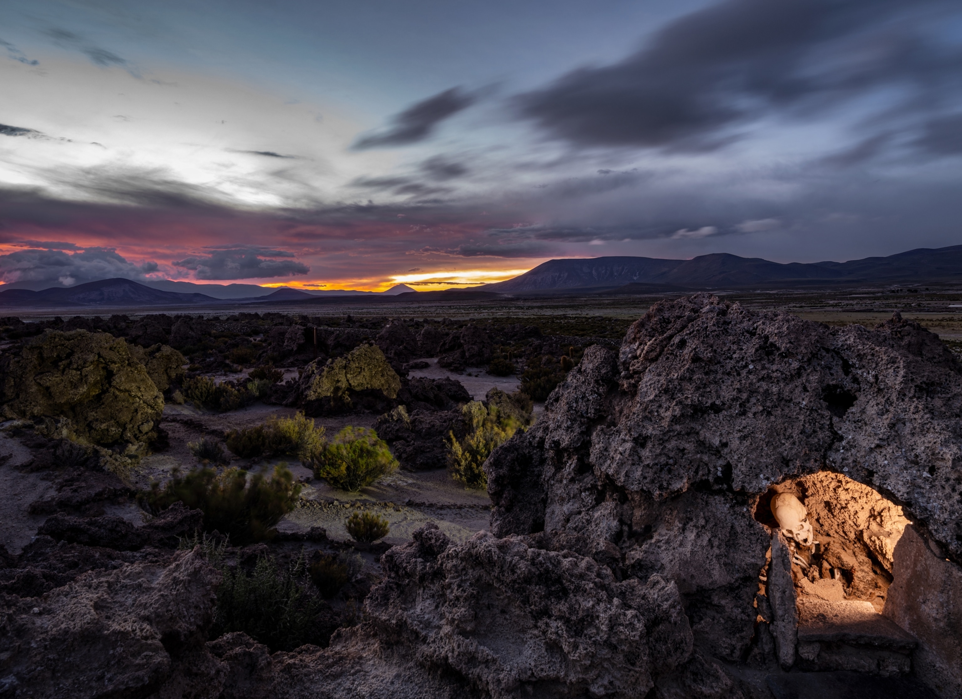 A vast landscape shows dirt, small and medium rocks, and small shrubbery spreading into the horizon. In the background are mountains and the sun setting behind them, creating a yellow, orange, and pink hue. In the foreground is a large rock with a small cave inside of it. Inside the is illuminated by a light, and visible is a human skull that appears to be sticking out from the wall.