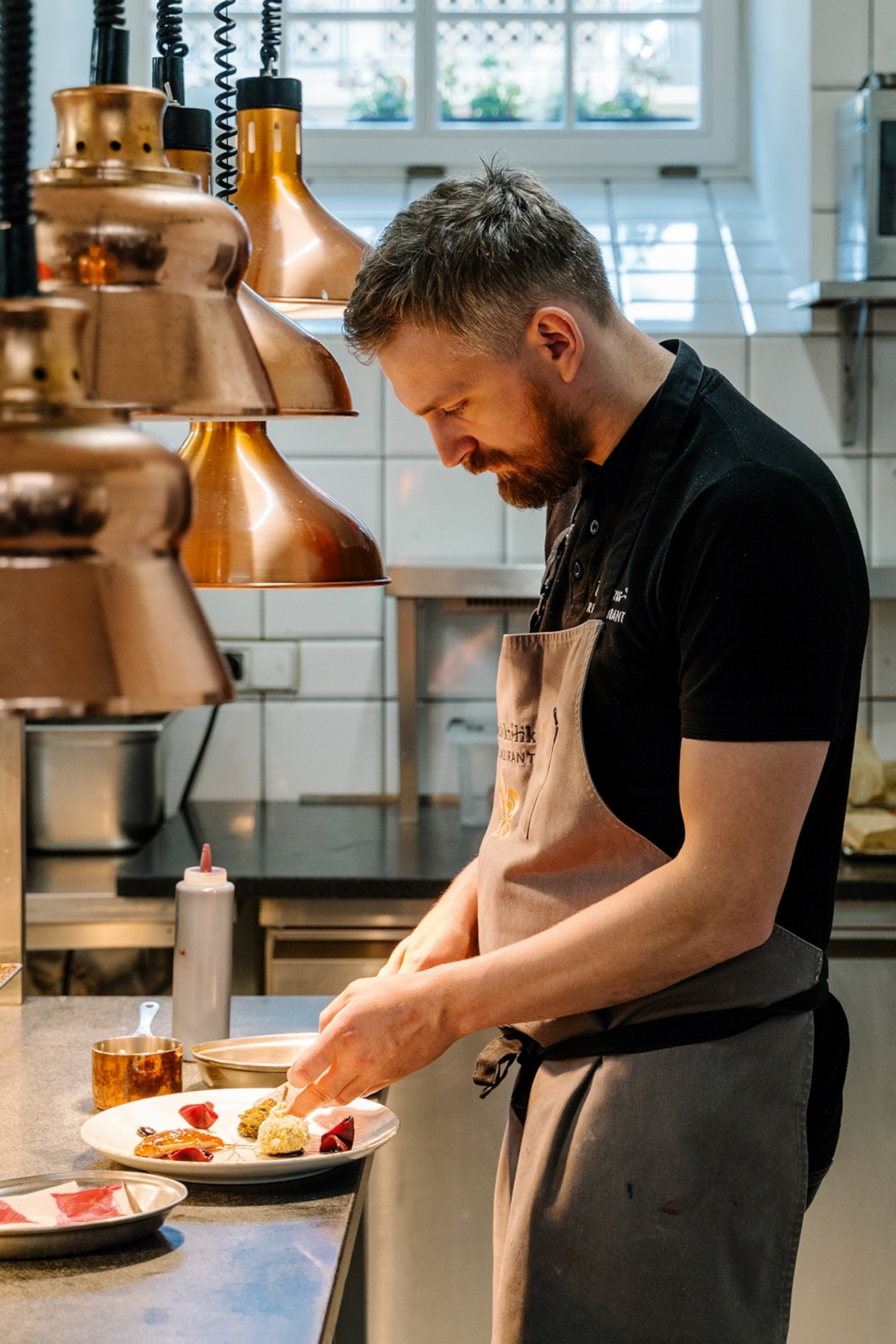 A young, bearded chef leaning over the kitchen pass in his restaurant, adding final touches to a couple of plated dishes.