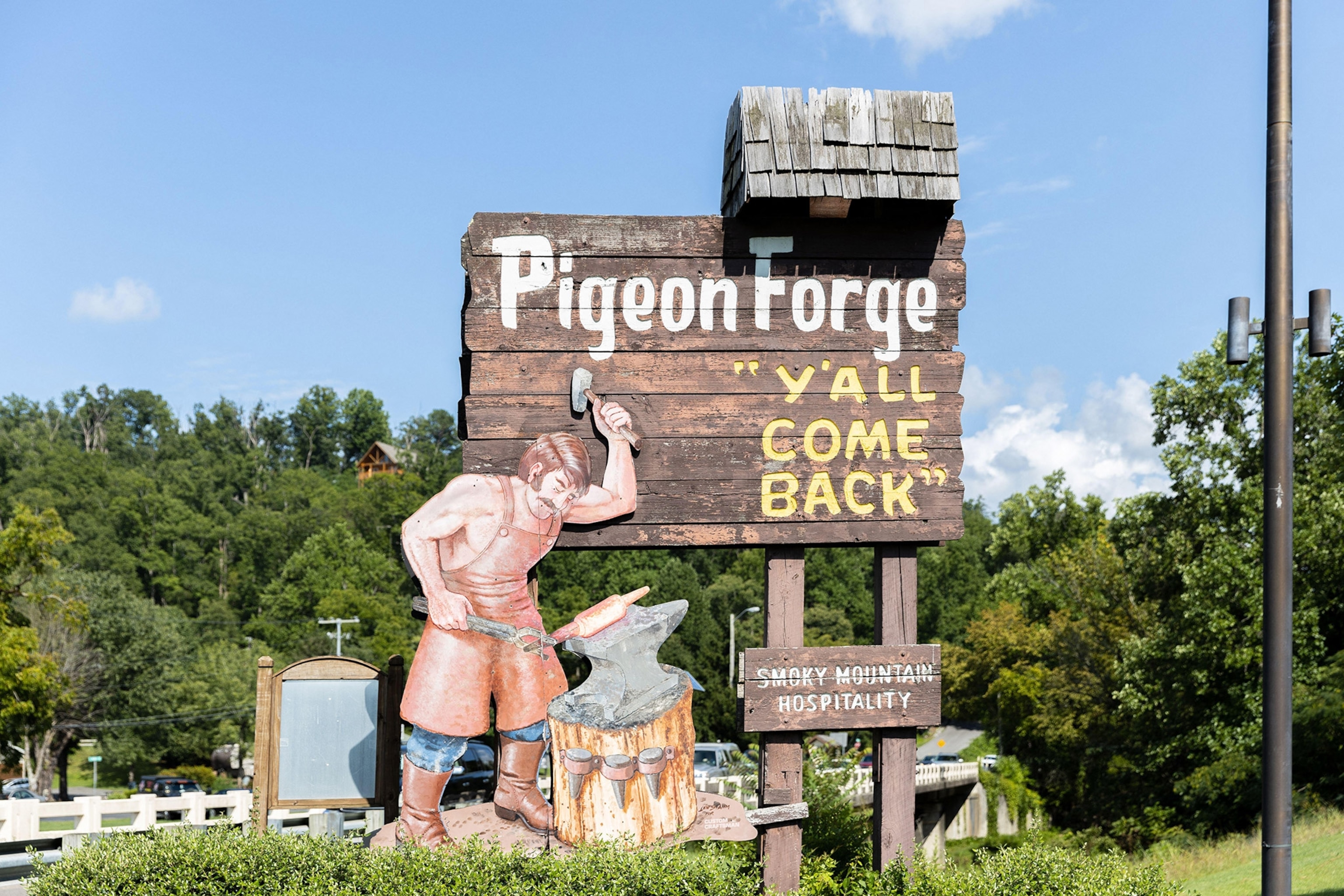 A large wooden sign reads "PIGEON FORGE, Y'ALL COME BACK" and shows an illustration of a blacksmith hammering hot steel. Trees surrounding.