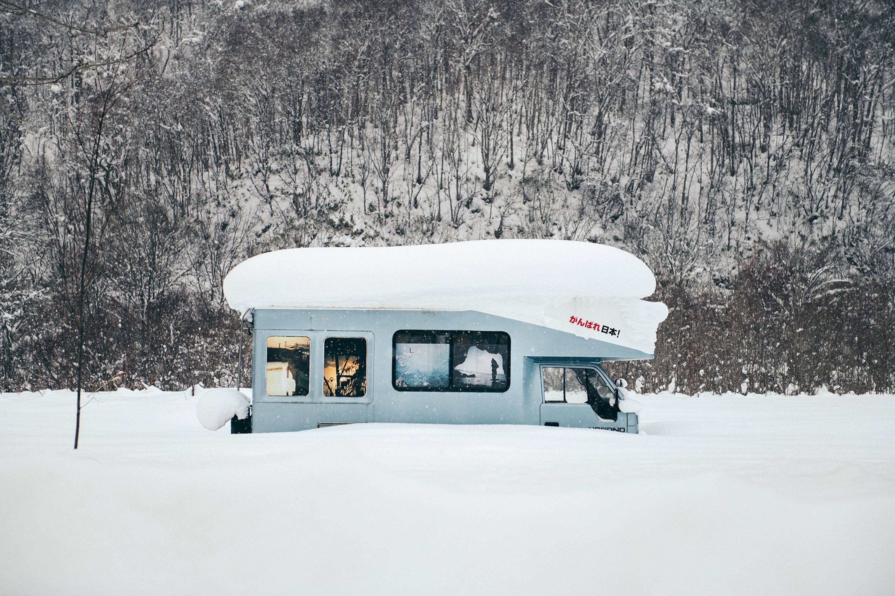 A campervan bus buried deep under a layer of powdery snow in a winter forest landscape.