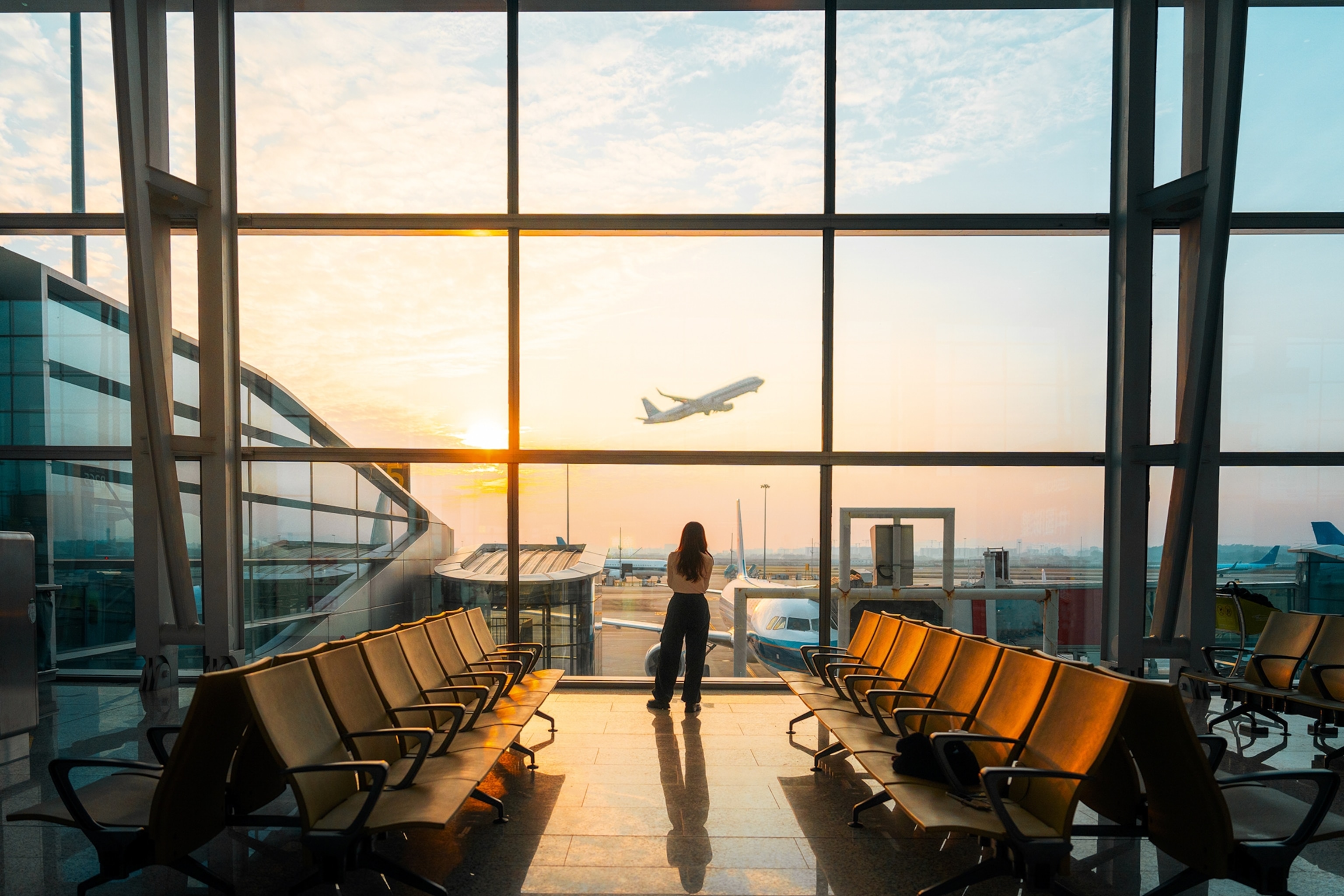 A person standing at an airport gate looking out through a glass front as a plane takes off.