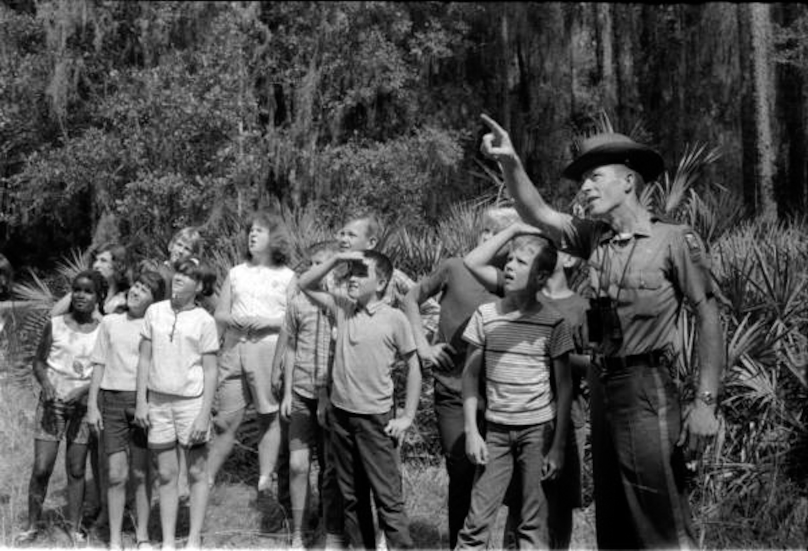 A black and white photograph of a man pointing with a group of children listening.