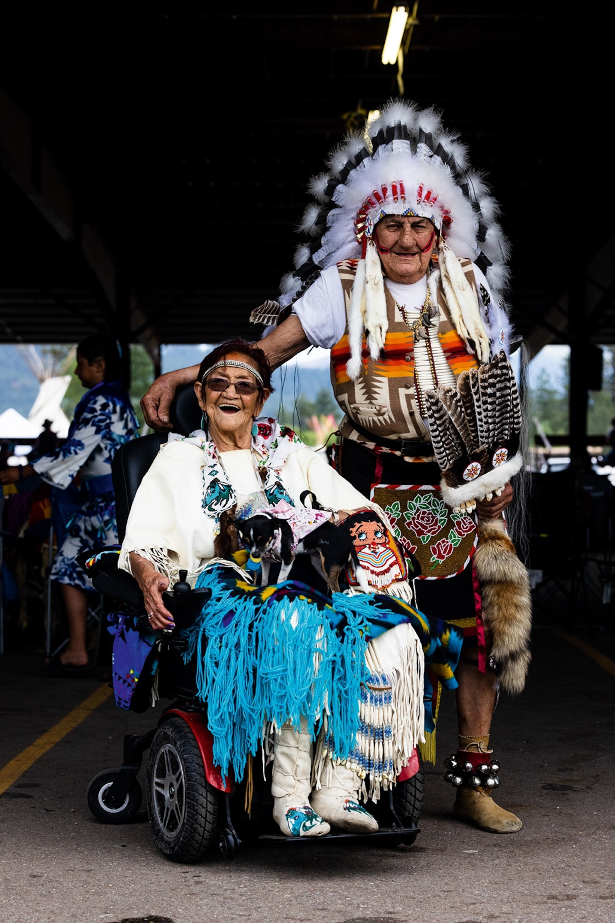 An elderly Native American couple - the woman in a wheelchair - posing for a photo in celebratory dress, like feathered head pieces and bells around their boots.