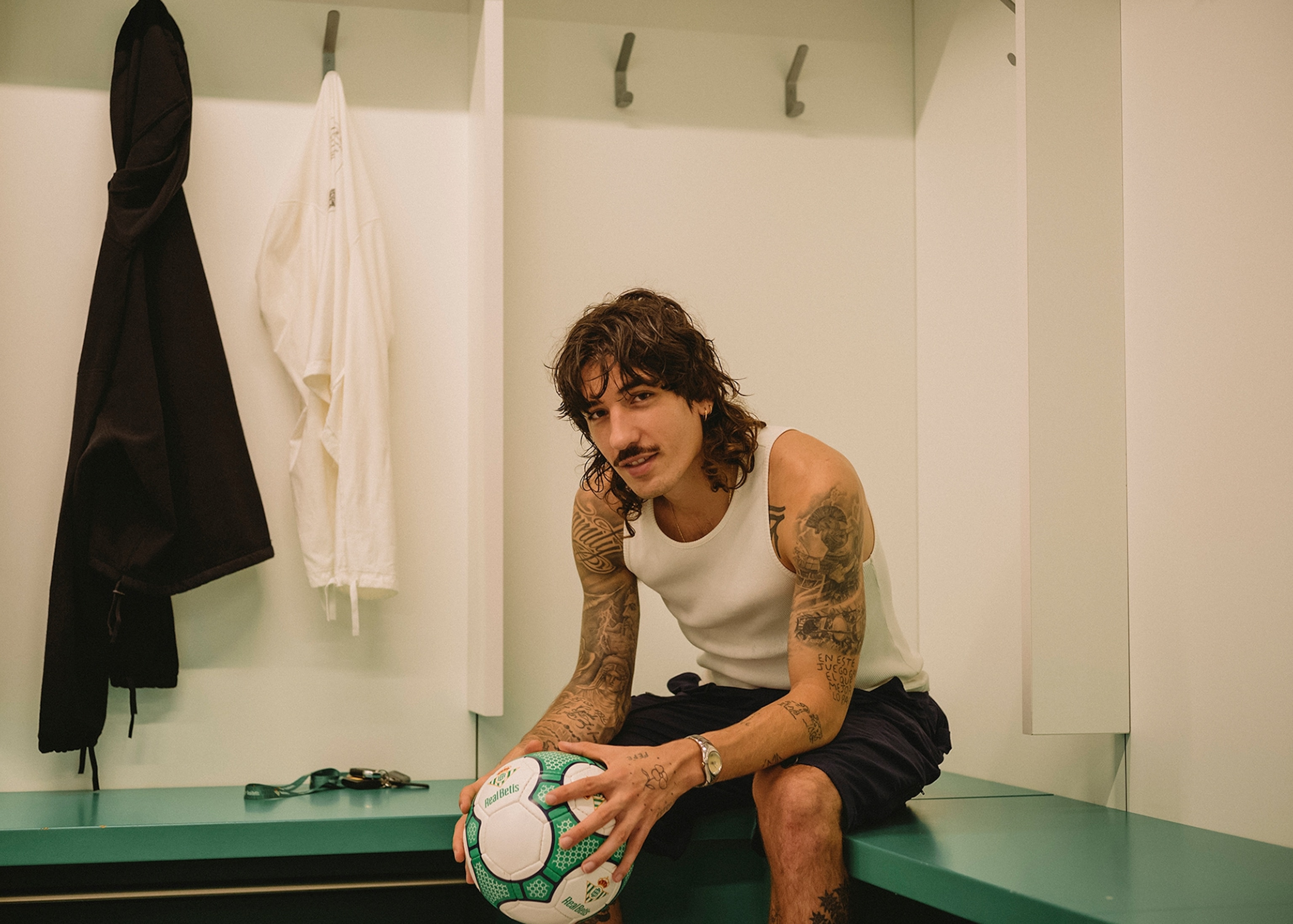 A man sits inside of a locker room on a green bench. He is holding a soccer ball.