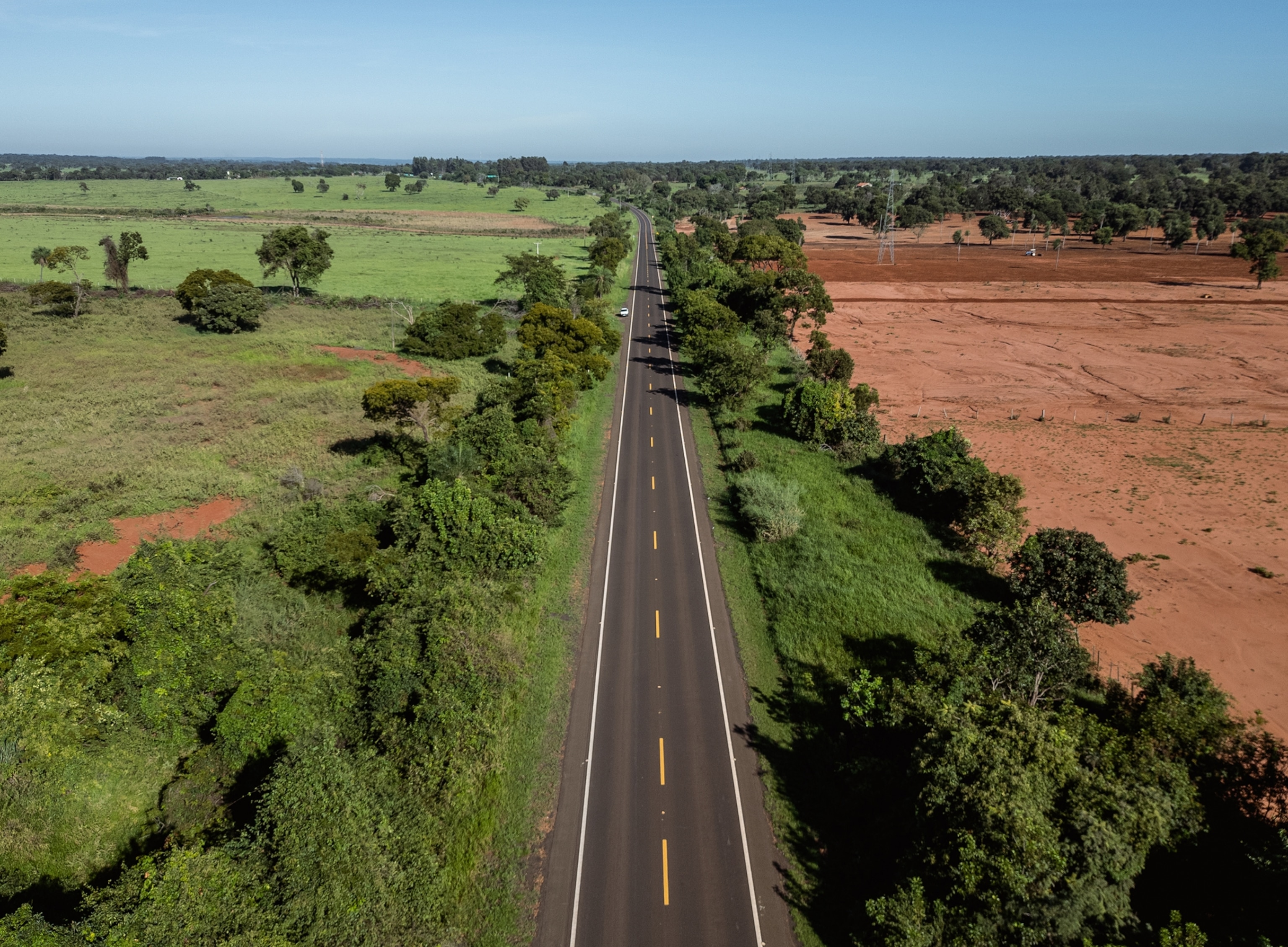 An aerial view of a highway that starts center frame in the foreground and stretches into the distance in the background. To the left of the highway is a large grass field with trees lining directly next to highway. To the right of the highway are trees lining directly next to the road and a red, dirt field with trees further off into the distance.
