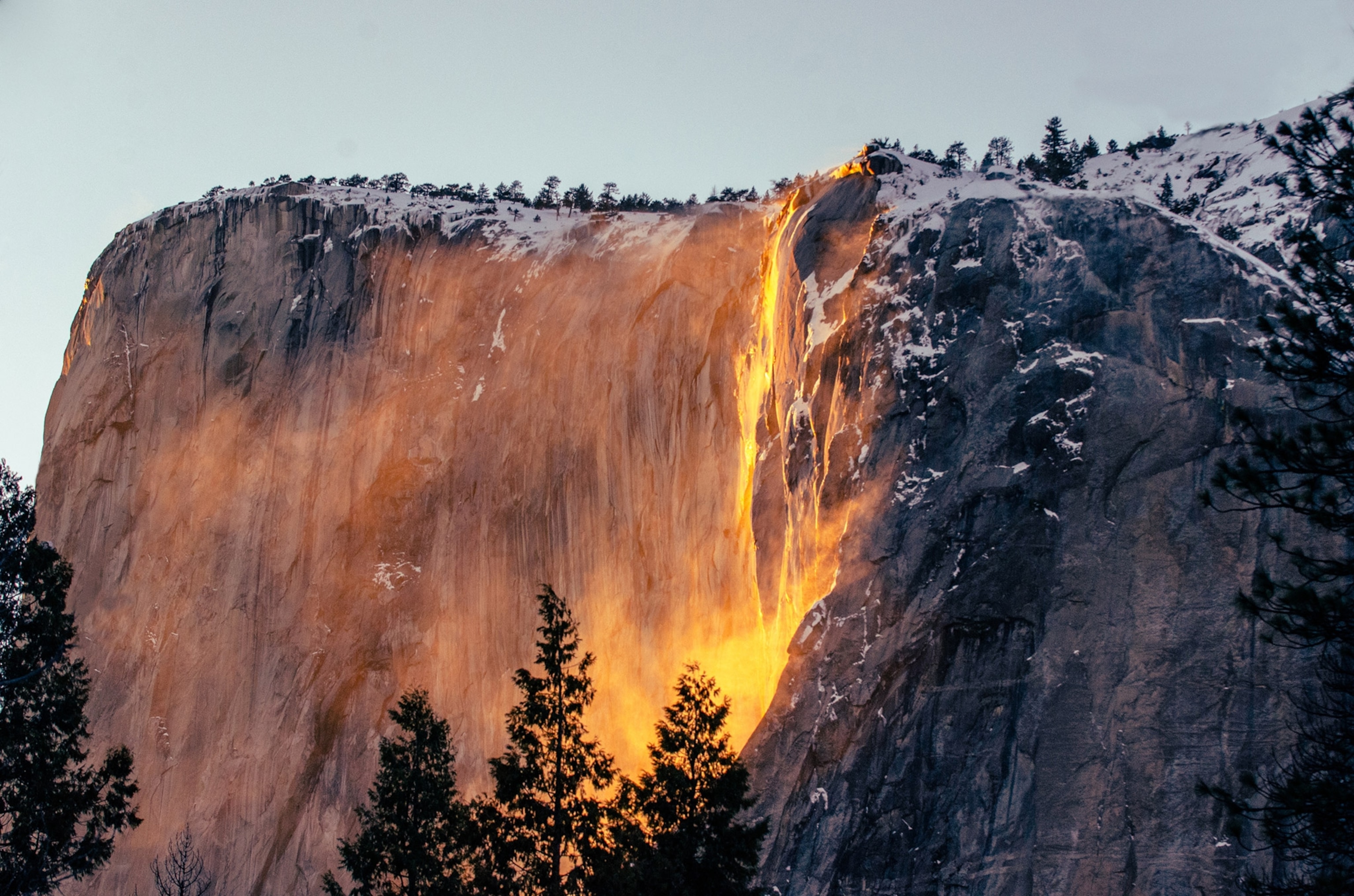 A glowing orange waterfall cascades down a steep granite face, capped by snow and surrounded by pine trees in foreground
