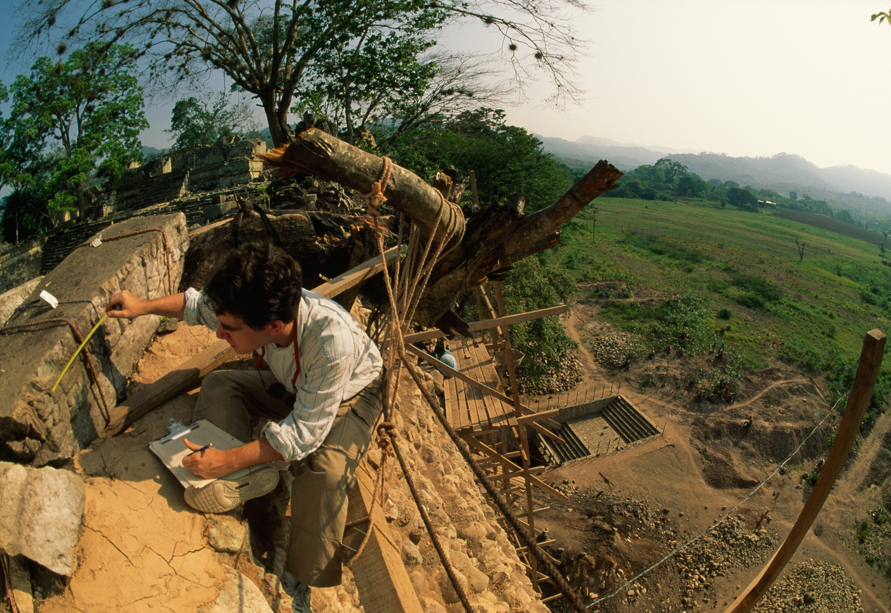 Archaeologist measuring ancient stone ruins on a steep site, surrounded by lush trees and overlooking vast green fields under a bright sky.