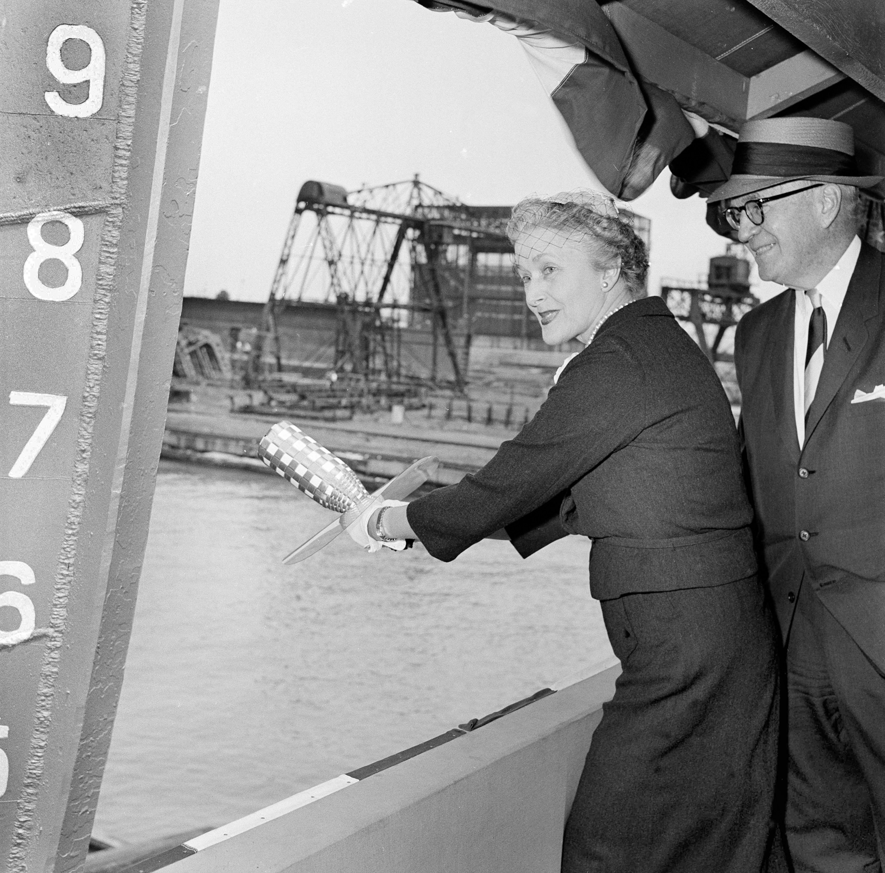 Elizabeth Fitgerald swinging a ceremonial bottle of champagne to launch the Edmund Fitzgerald as her husband looks on