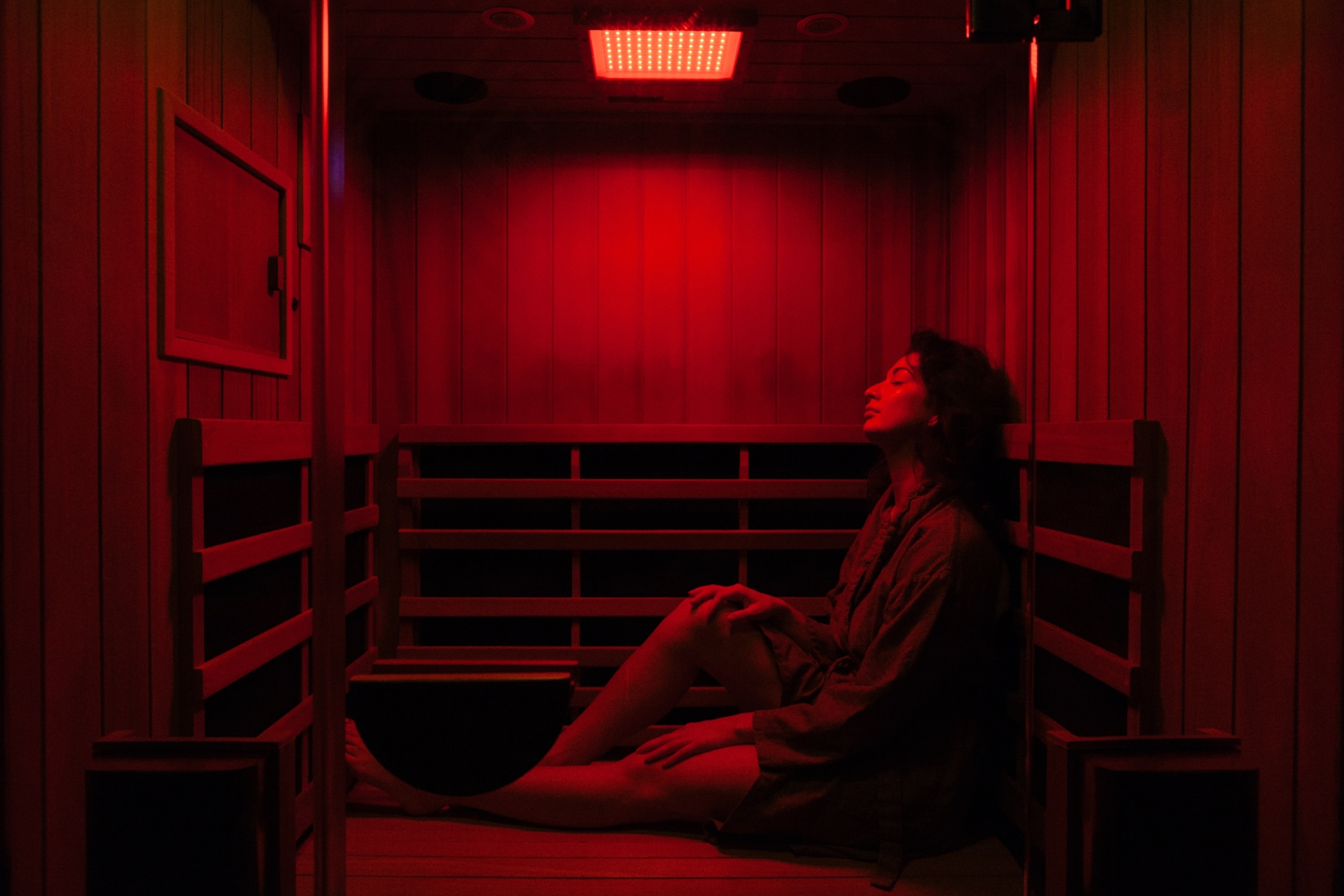 A woman relaxing in a dimly lit red infrared sauna room, sitting with eyes closed, wearing casual clothing